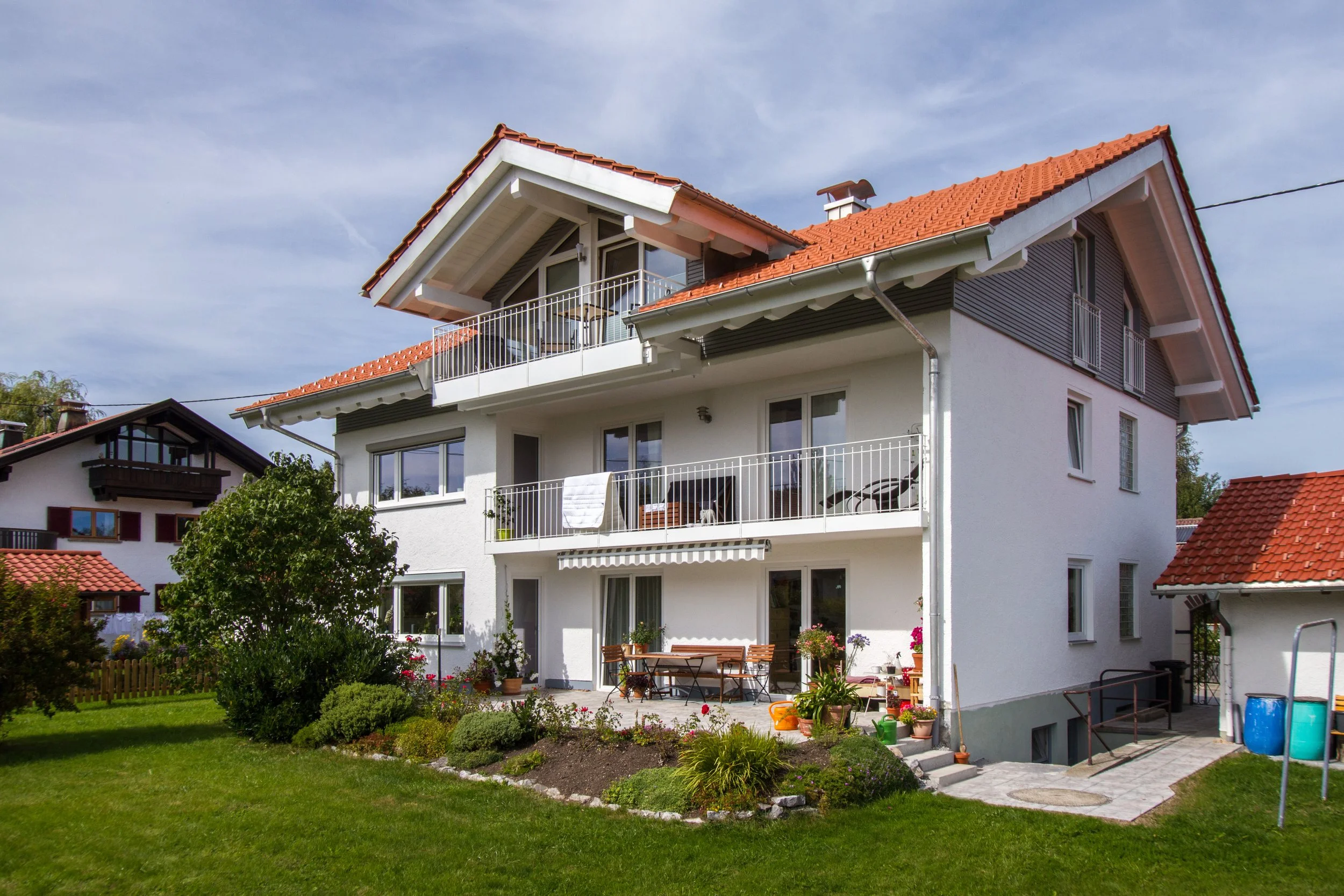 Ein mehrstöckiges Haus mit weißen Wänden, rotem Ziegeldach und Balkon im obersten Stockwerk. Im Garten sind Blumen, Büsche und eine Terrasse mit Gartentisch und Stühlen.