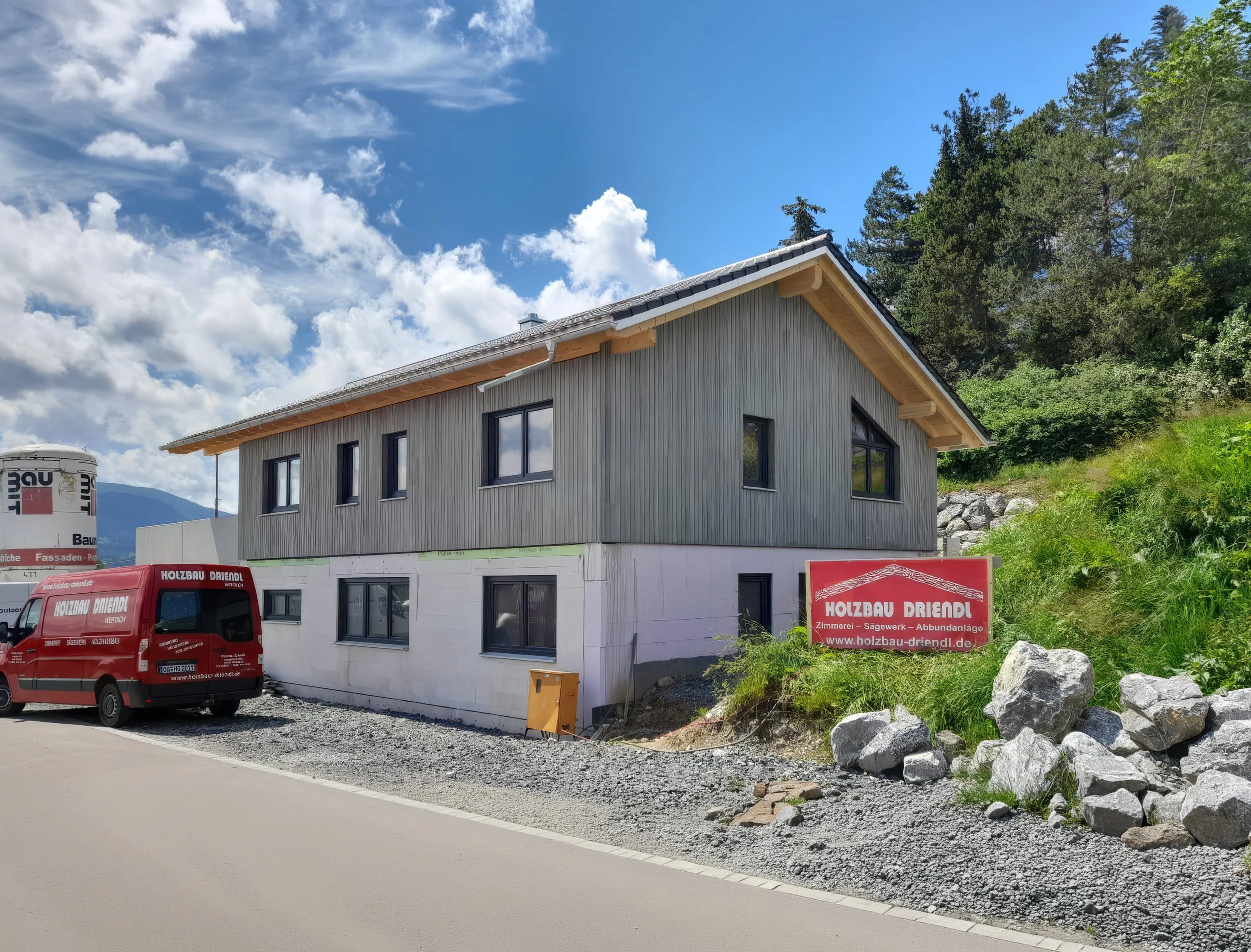 Bauhaus-Fahrzeug und Baustellenschilder vor einem Haus im Bau, mit Wald im Hintergrund, blauer Himmel und Wolken.