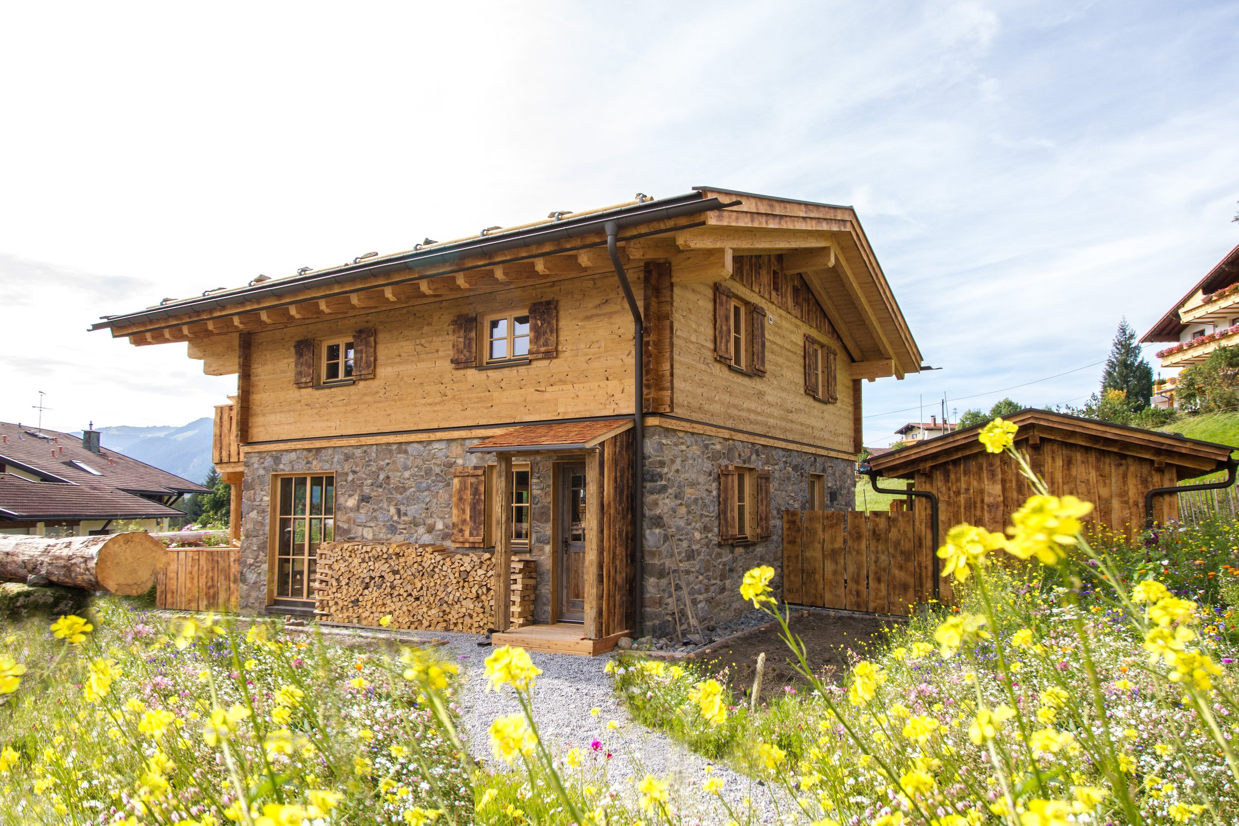 Holzhaus mit Steinfundament, im Garten mit bunten Blumen und einem Holzstapel im Vordergrund.