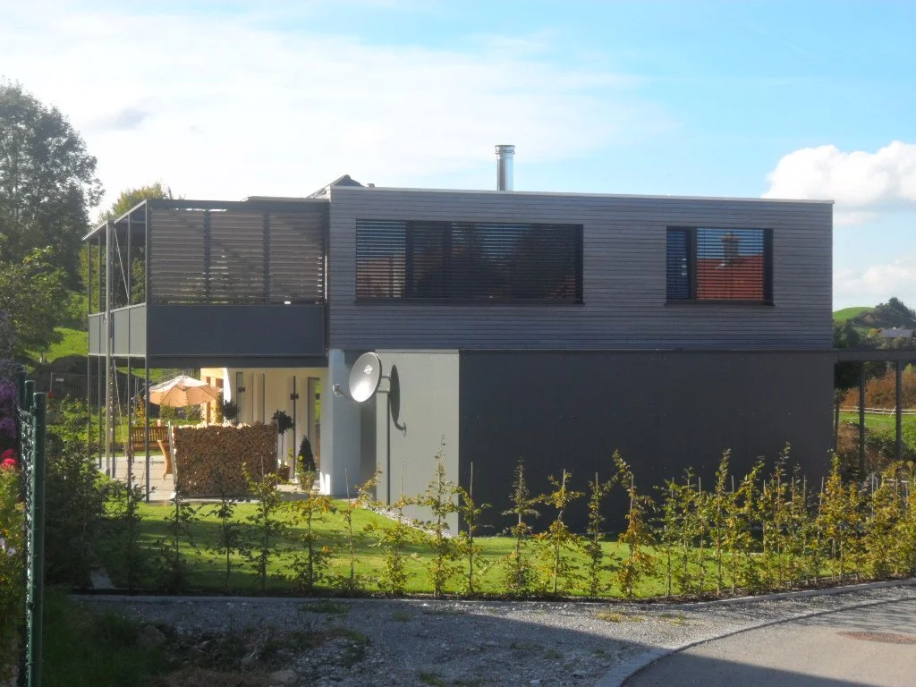 Modernes zweistöckiges Haus mit dunklem oberen und hellen unteren Etagen, umgeben von Garten und Bäumen, bei sonnigem Himmel.