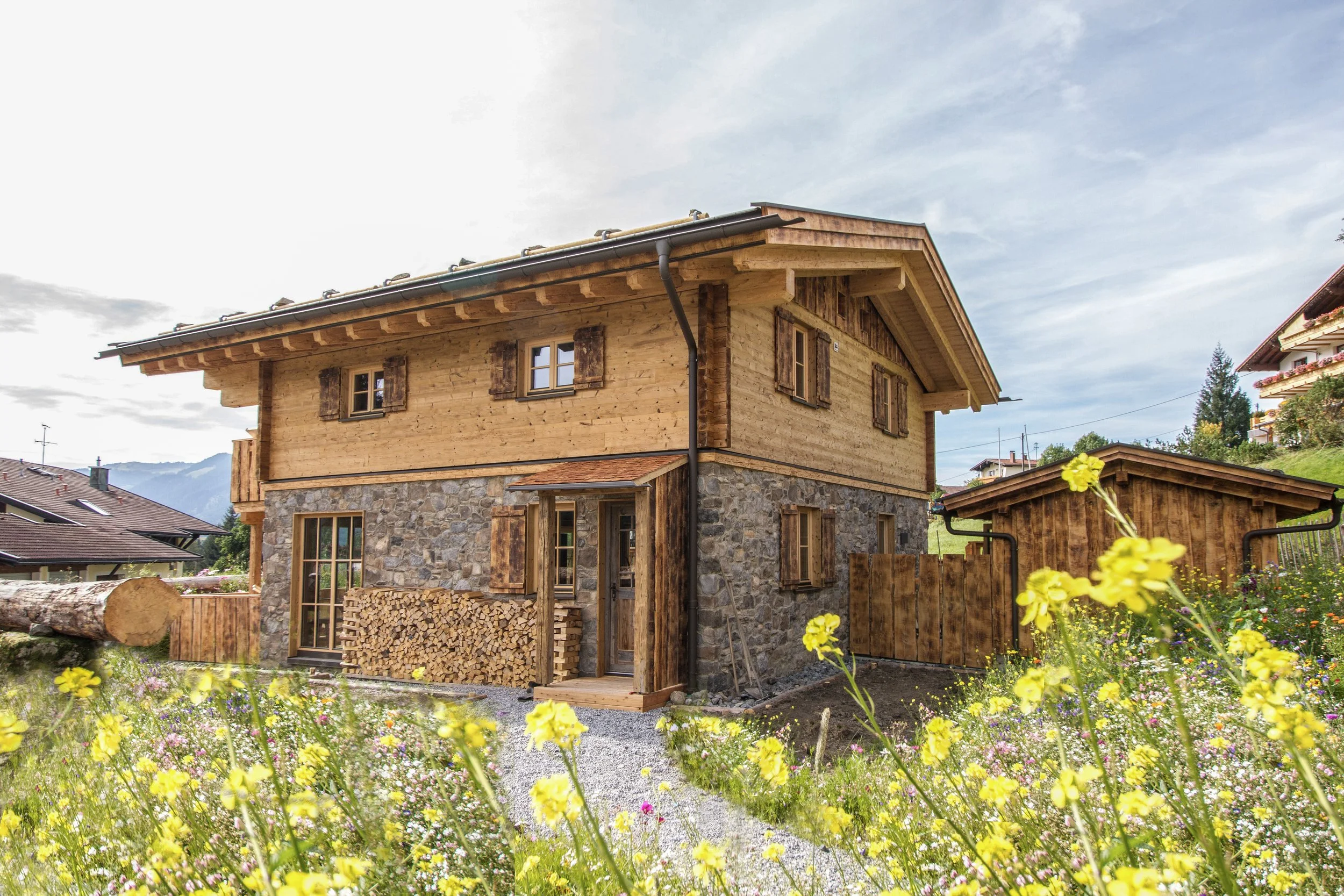 Ein zweistöckiges Haus im Chaletstil mit Holz und Stein im ländlichen Bereich, umgeben von bunten Blumen und einem Garten, mit bewölktem Himmel im Hintergrund.