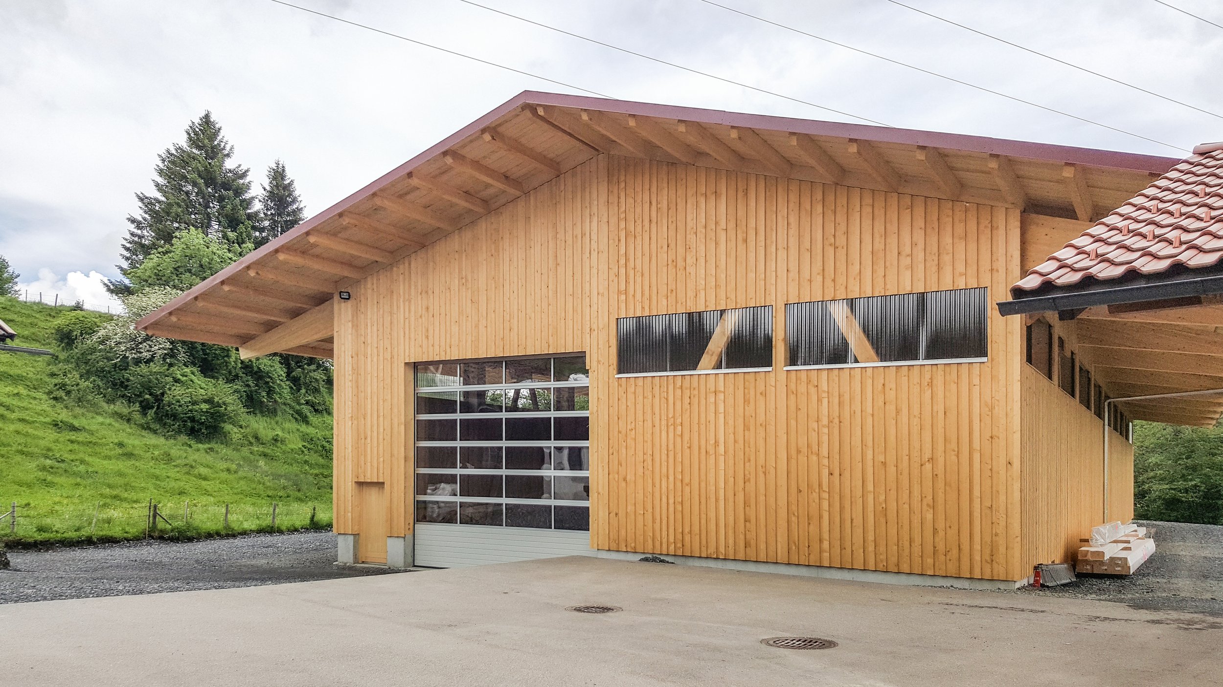 Holzbau mit großen Garage-Toren und modernem Design, umgeben von grüner Landschaft, im Hintergrund Bäume und bewölkter Himmel.