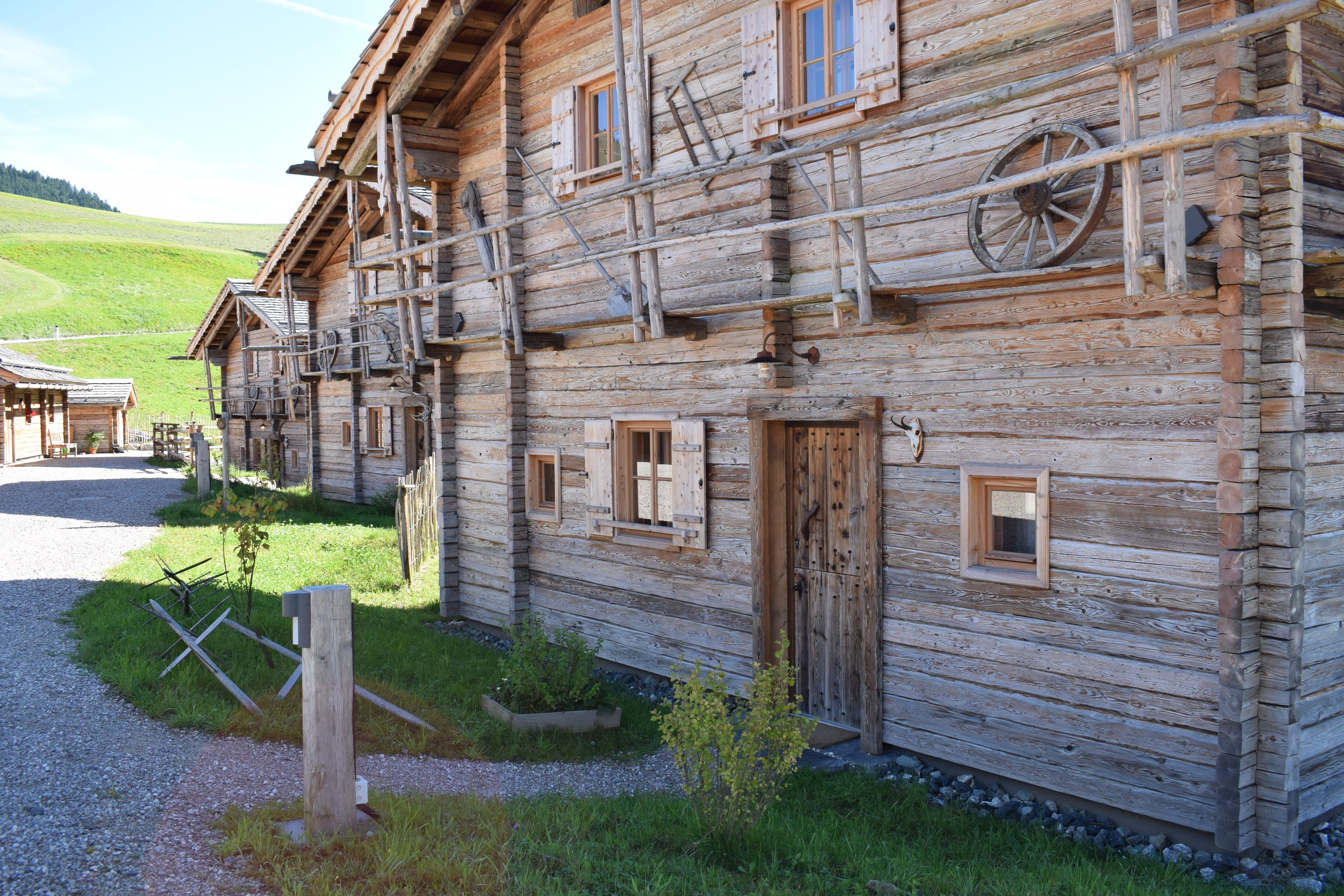 Ein Holzhaus mit mehreren kleinen Fenstern und Holzschindeln im ländlichen Gebiet, umgeben von grünen Wiesen und Bergen im Hintergrund.