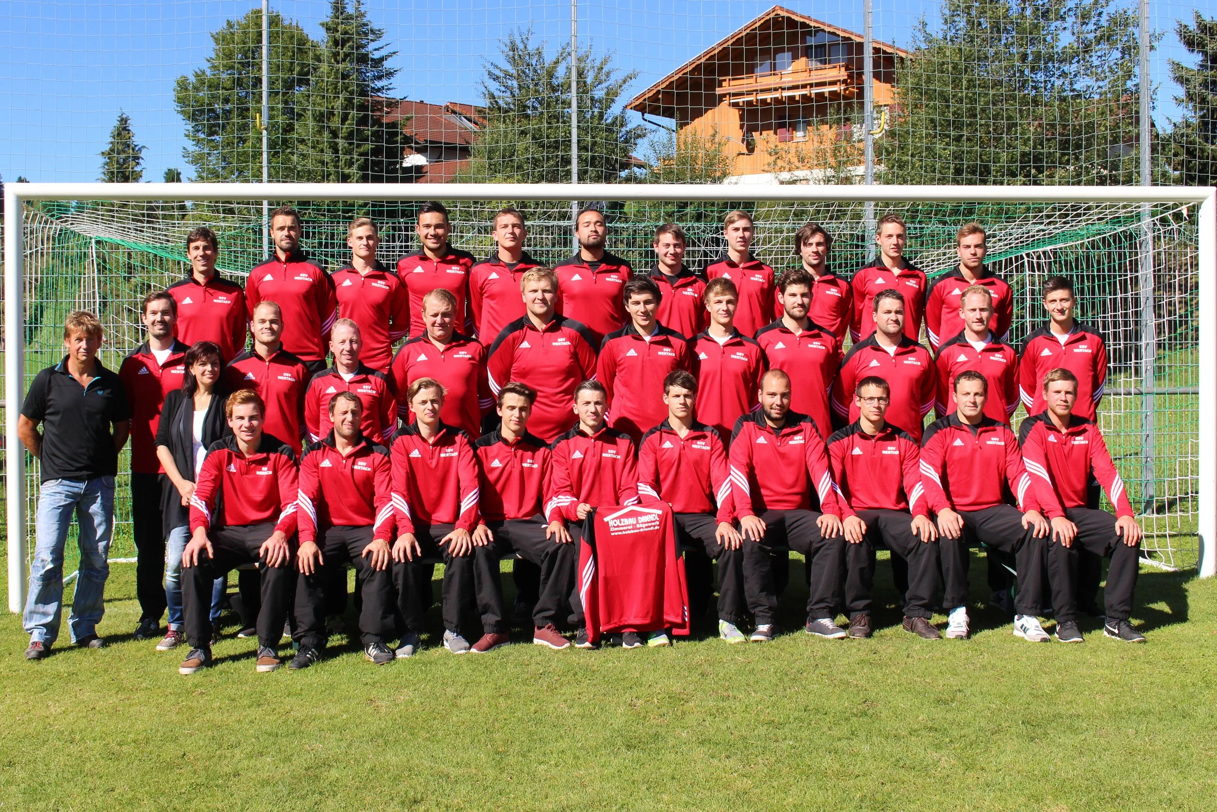 Gruppenfoto eines Fußballteams auf einem Spielplatz, alle tragen rote Trikots, im Hintergrund ist ein Fußballtor und ein Haus im Grünen.