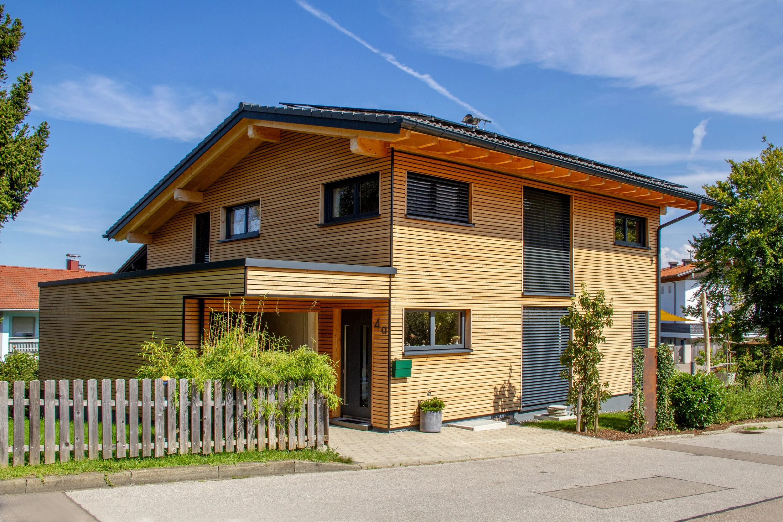 Moderner zweistöckiger Holzhaus mit schwarzem Dach, großen Fenstern und Garten, umgeben von Bäumen und Zaun, bei sonnigem Himmel.