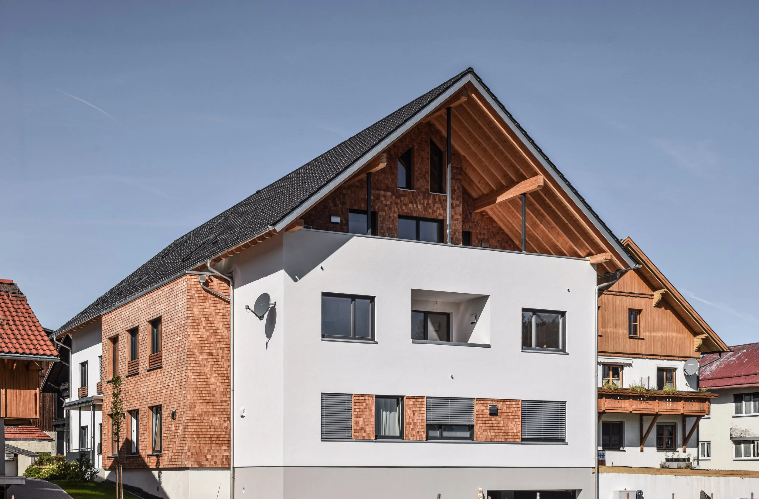 Modernes mehrstöckiges Haus mit weißen und roten Ziegelwänden, großen Fenstern, einem Balkon und einem Satteldach aus Schindeln, under dem blauer Himmel