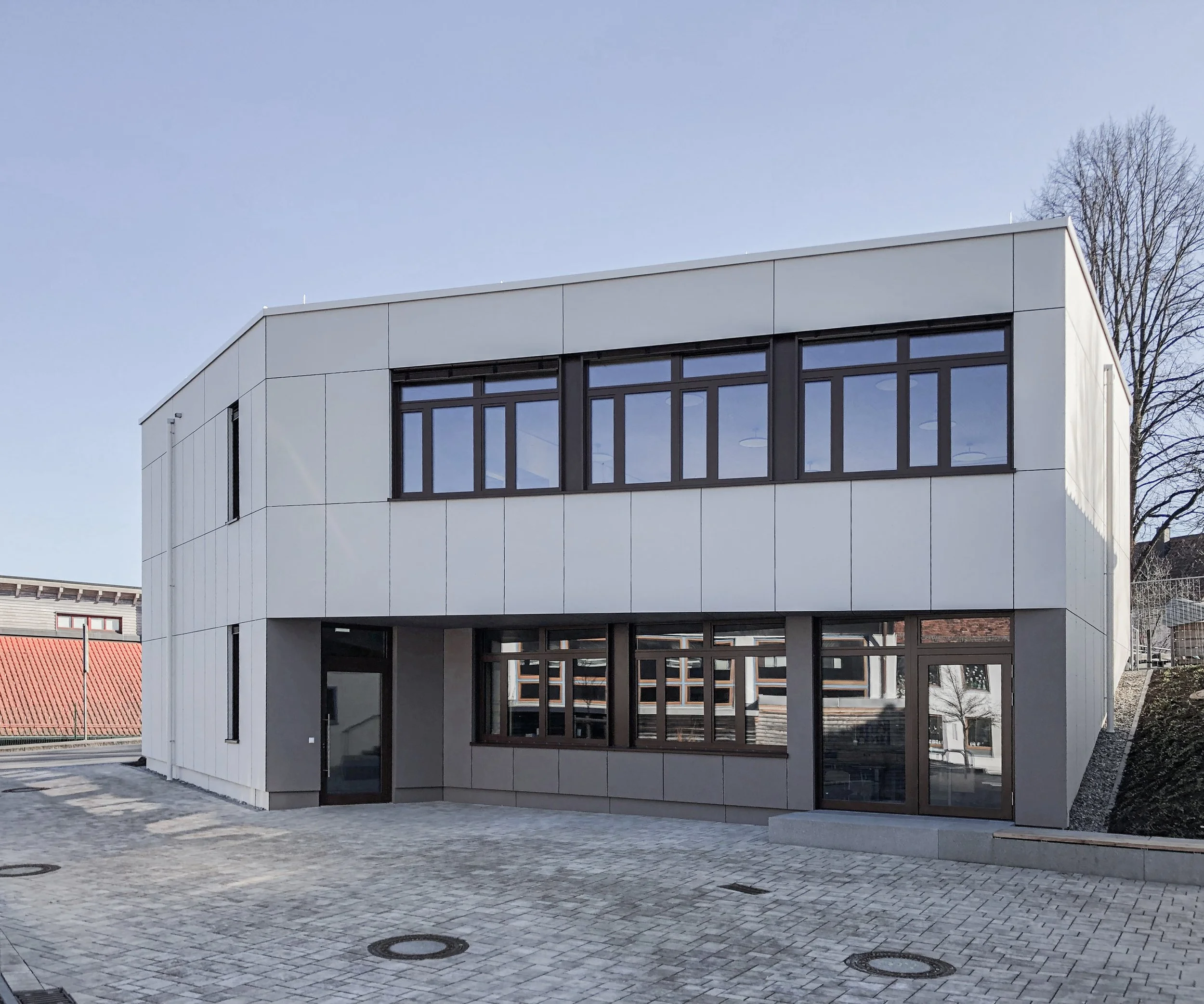 Moderne weiße Gebäudefassade mit großen Fenstern auf einer gepflasterten Fläche. Im Hintergrund sind vereinzelte Bäume und ein klarer Himmel sichtbar.