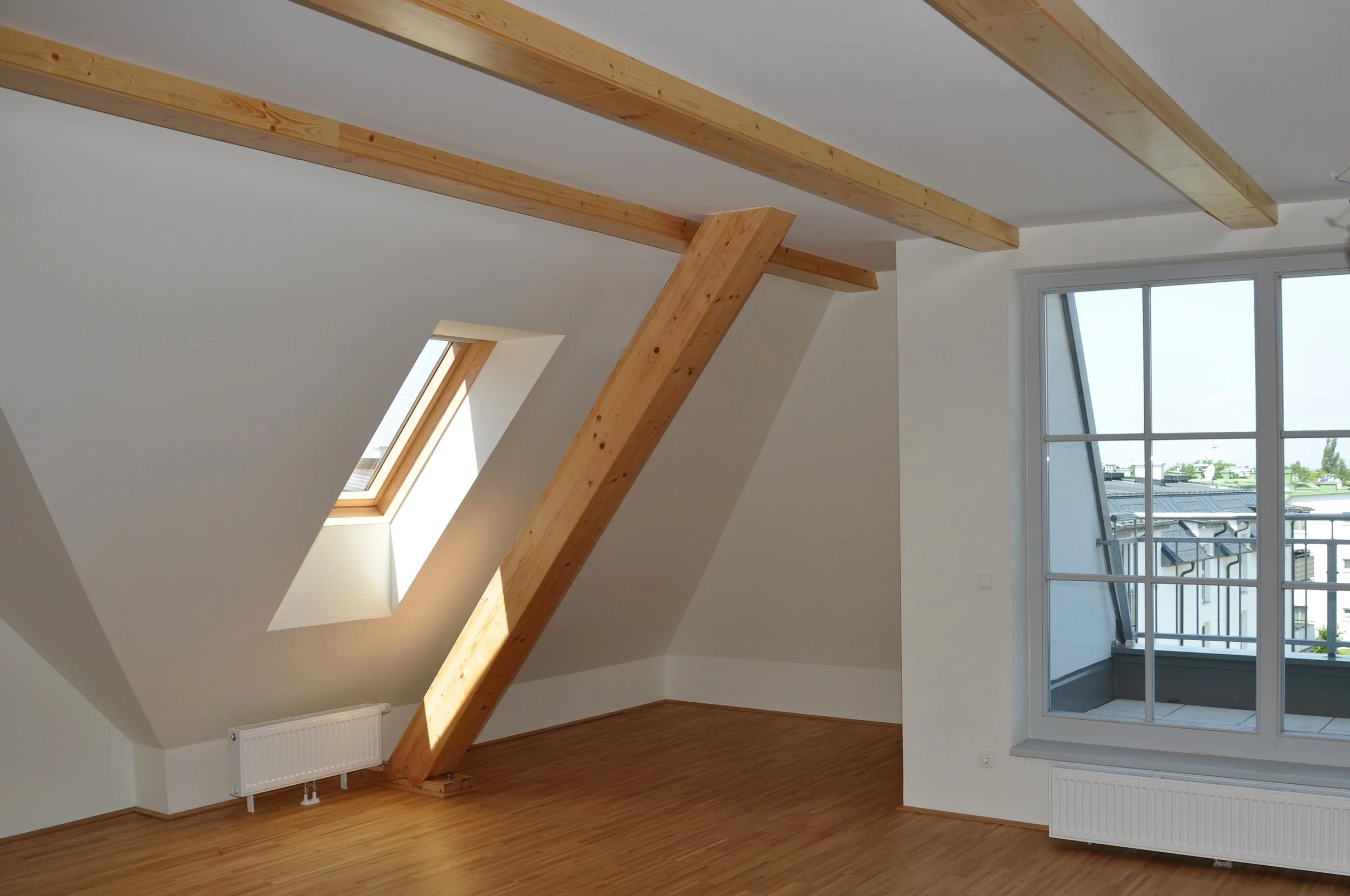 Ein leerer Raum mit weißen Wänden, Holzboden und sichtbaren Holzbalken an der Decke. Es gibt eine große Fensterfront, die auf einen Balkon hinausführt, und Dachfenster auf der schrägen Decke.