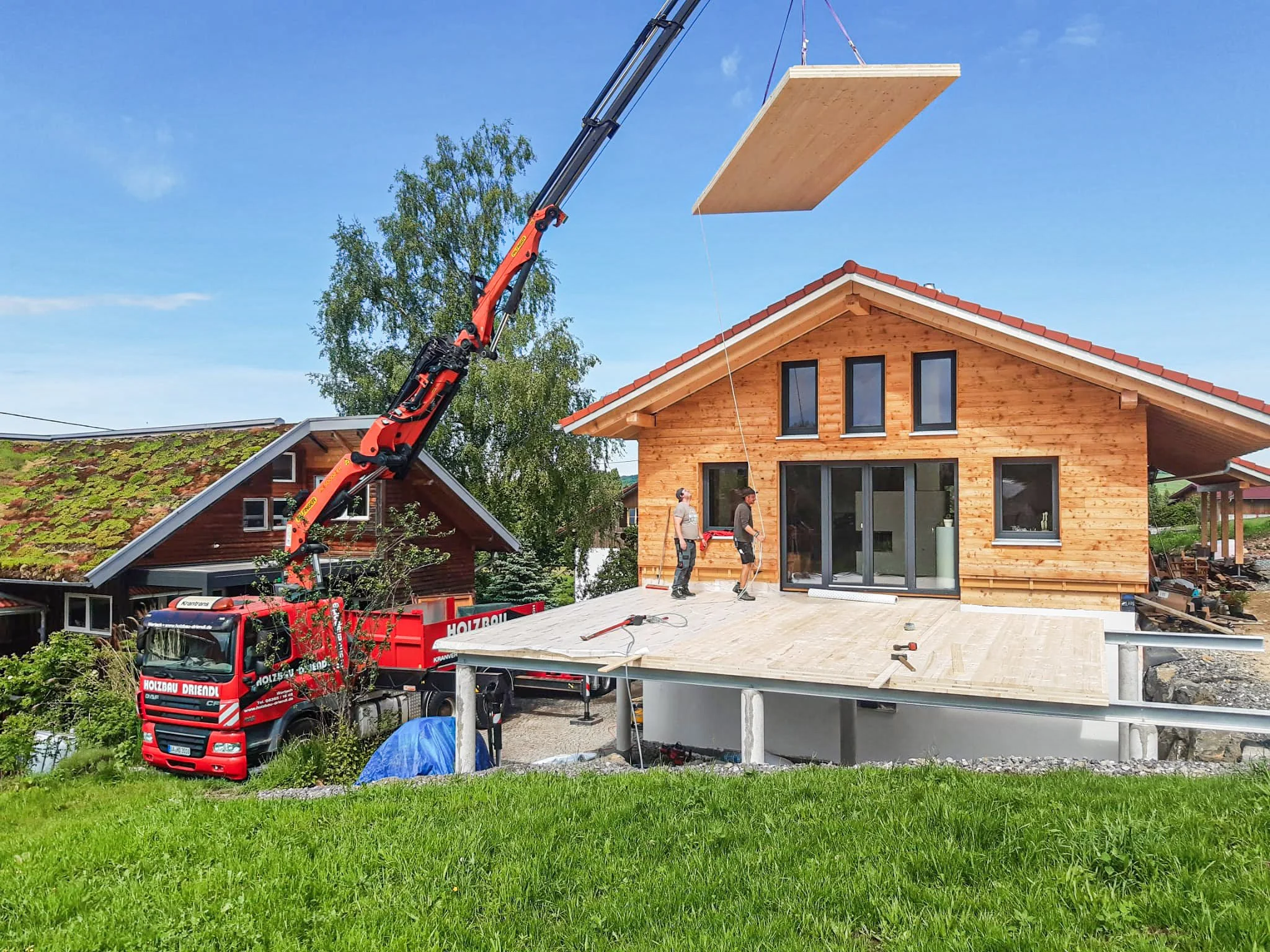 Ein Kran hebt eine große Holzplatte auf ein neues Holzhaus. Es sind drei Männer vor Ort, die den Bau beobachten.