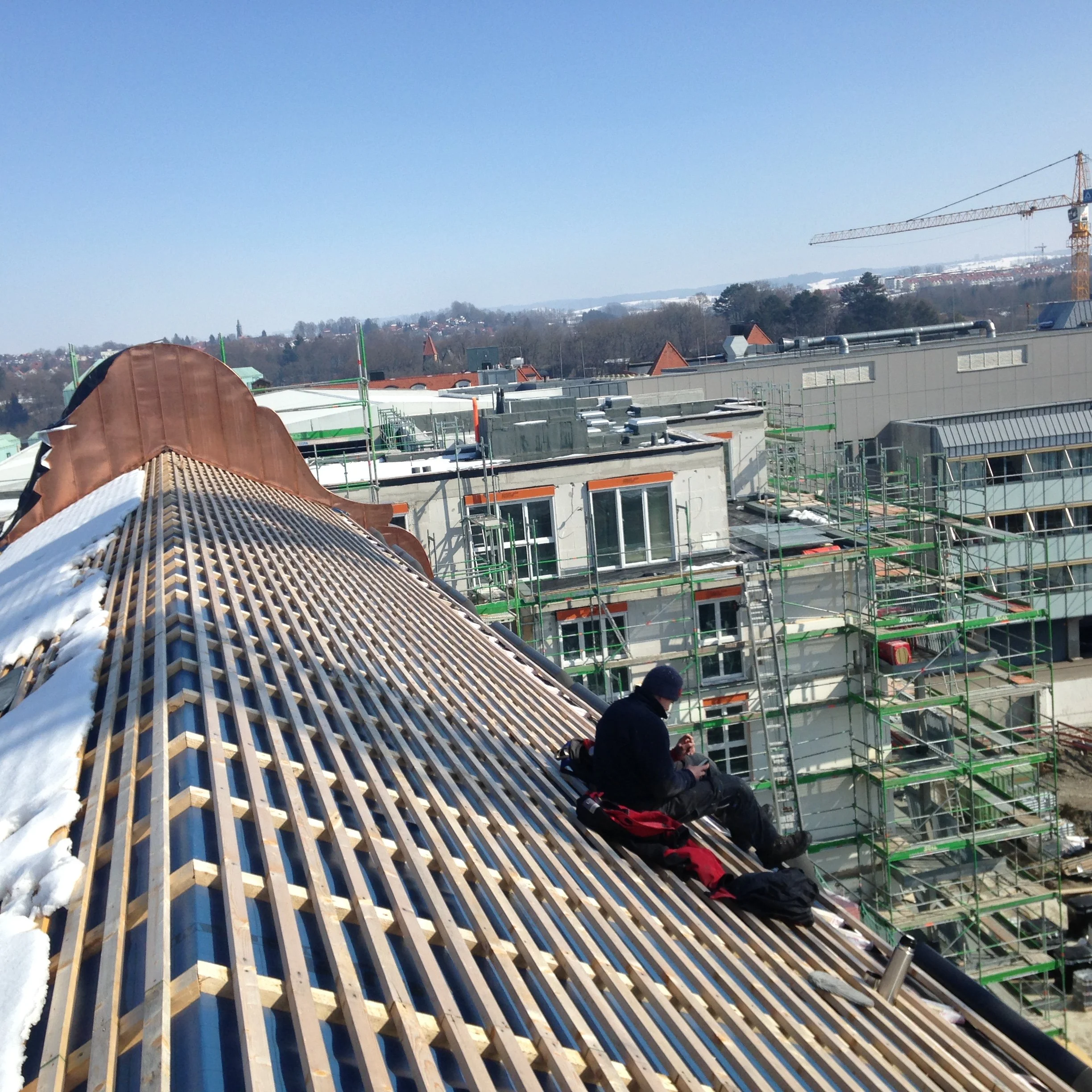Blick auf eine Baustelle, auf der ein Dach mit Holzrahmen gebaut wird, im Hintergrund Gebäude und ein Kran, unter einem klaren blauen Himmel.