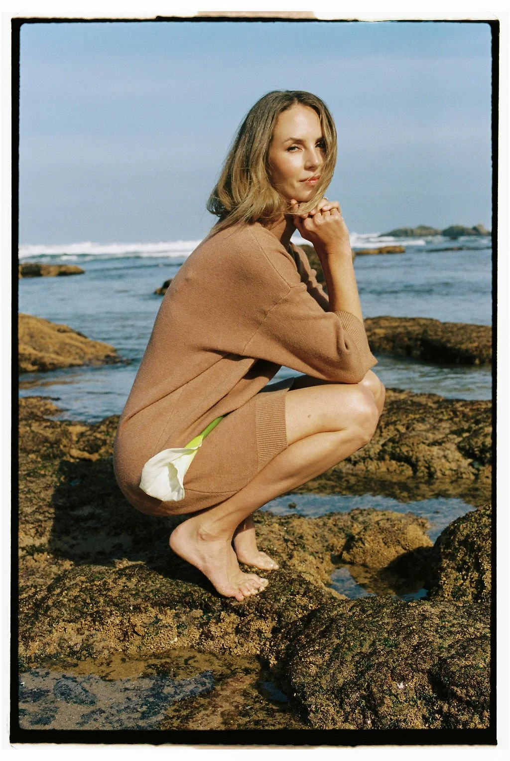 Keiarna from Gentle Currents crouching on rocks by the ocean, wearing a tan sweater and holding a white flower, with the sea and distant rocks in the background.