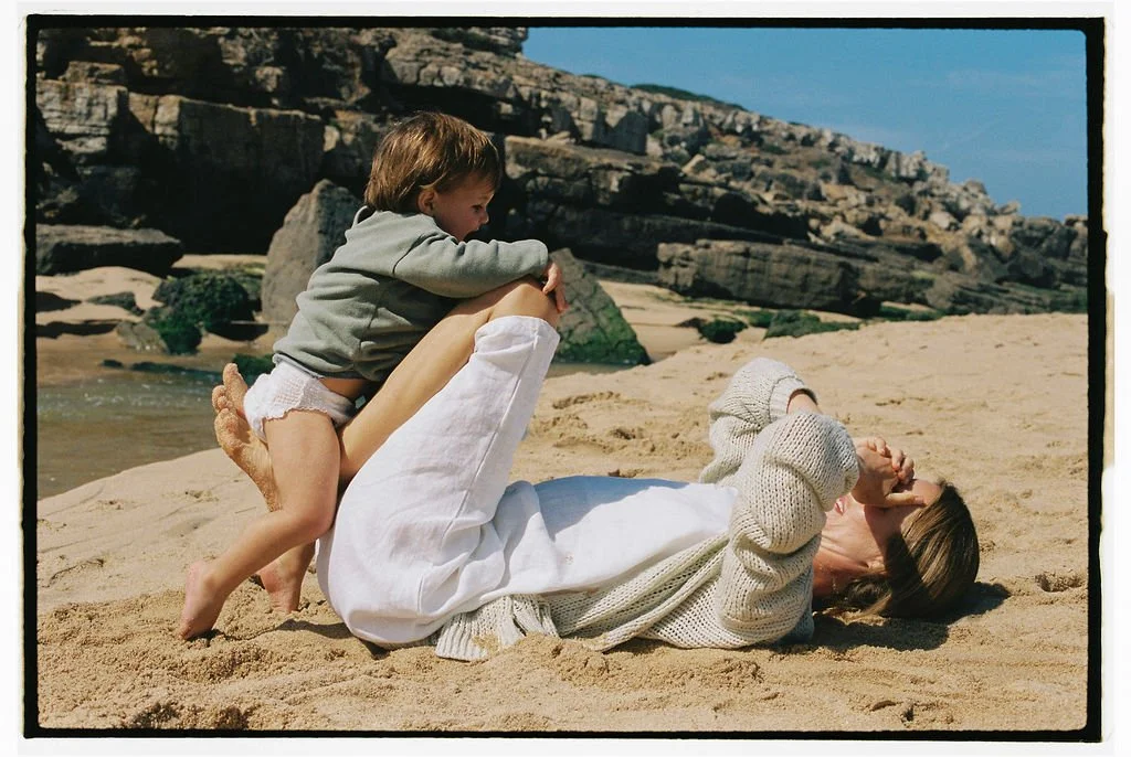 Keiarna from Gentle Currents and her young child playing on a sandy beach with rocks and cliffs in the background.
