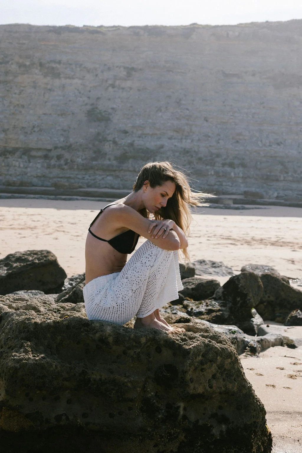 Keiarna from Gentle Currents sitting on rocks at the beach during sunset, wearing a black bikini top and white crocheted pants, looking down pensively.