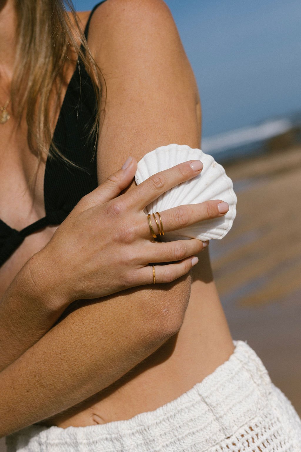 Person holding a white seashell on their arm at the beach, wearing gold rings and a black top.