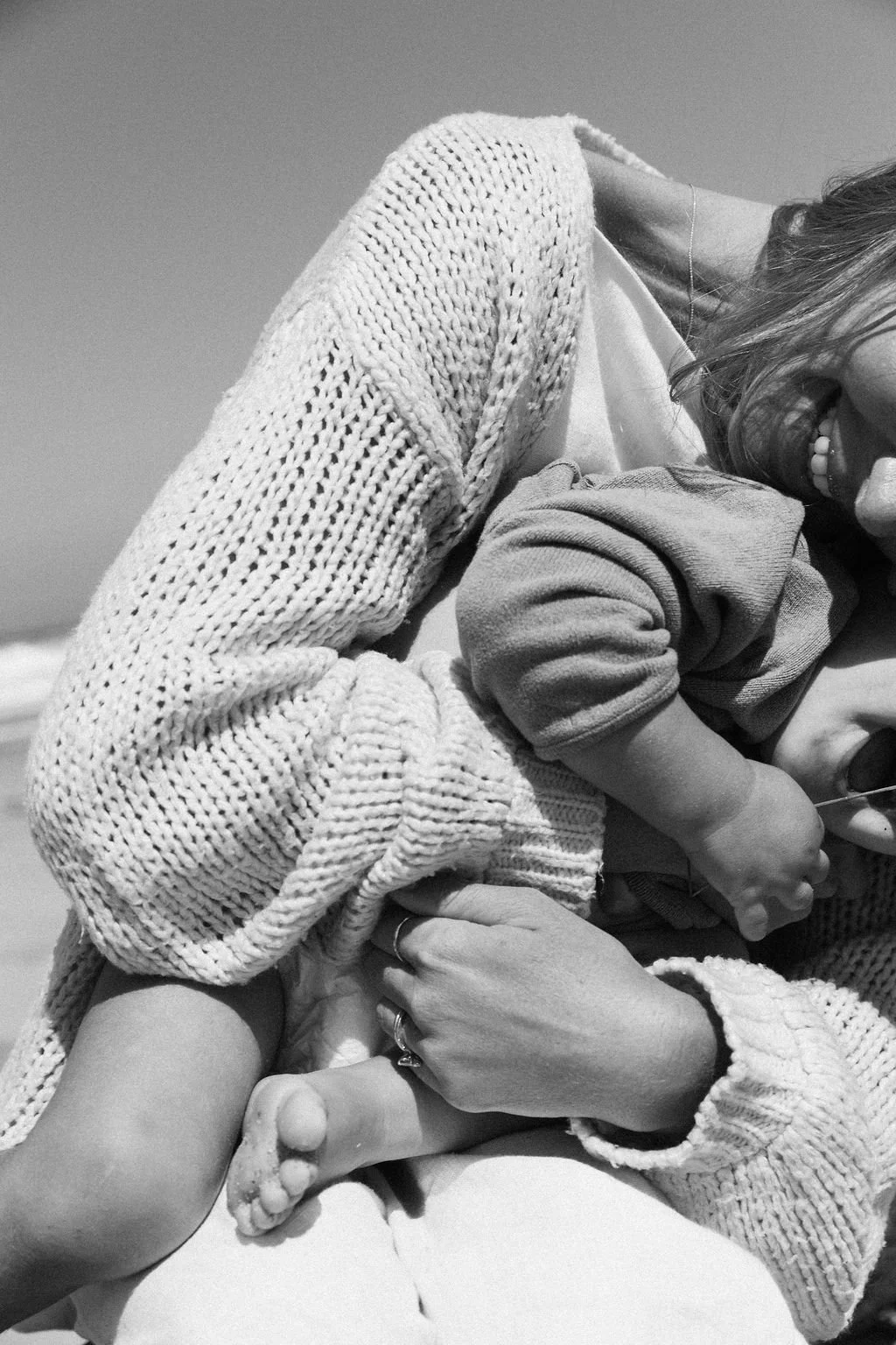 Keiarna from Gentle Currents holding her young child in their arms at the beach during the daytime, both wearing sweaters.