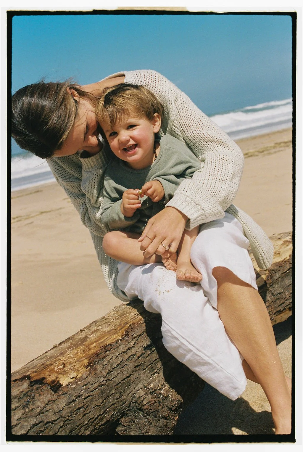 Keiarna from Gentle Currents and her young child sitting on a large piece of driftwood on the beach, smiling and laughing with the ocean and blue sky in the background.