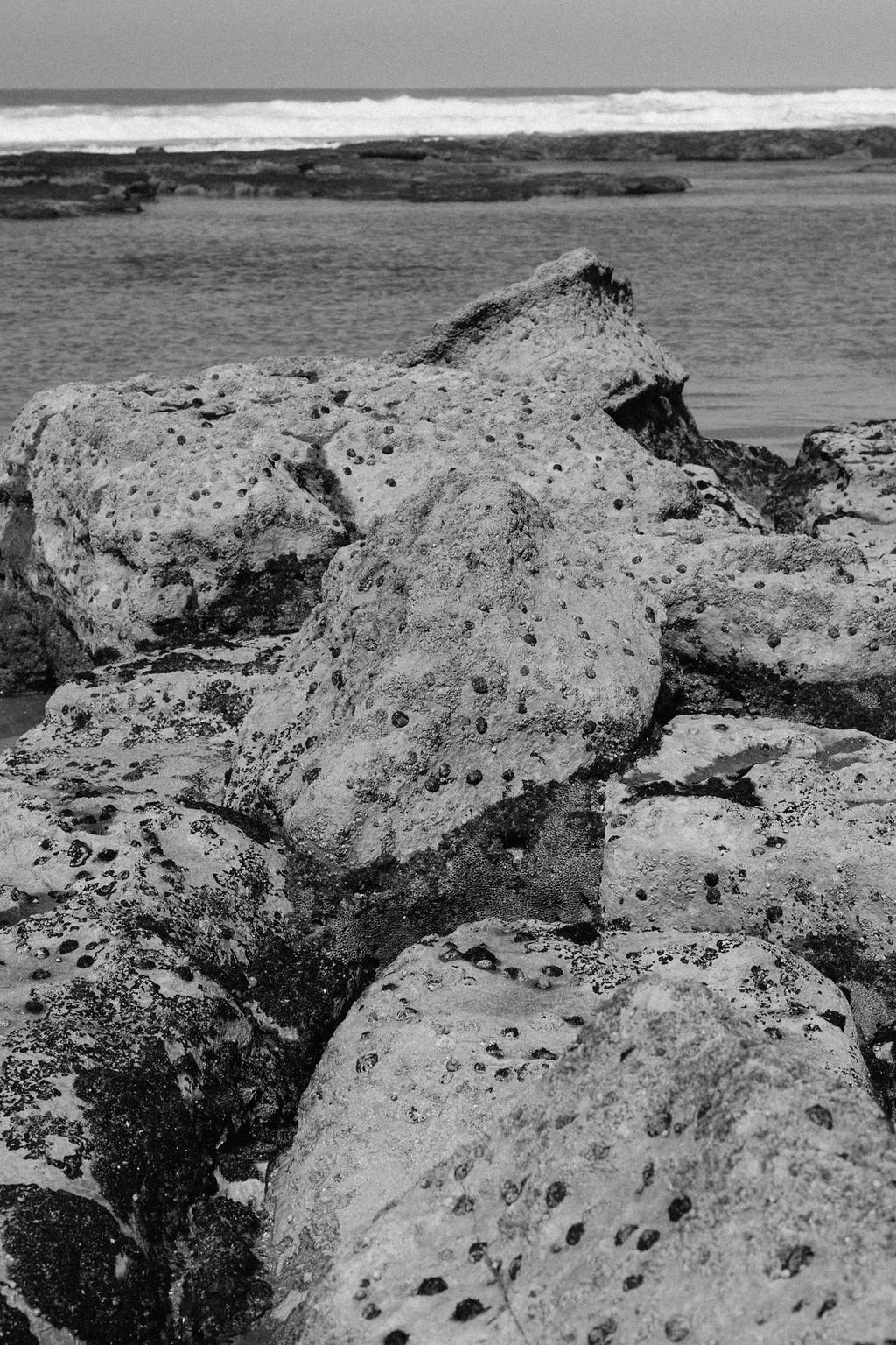 Black and white photo of large rocks along a beach with the ocean and waves in the background.