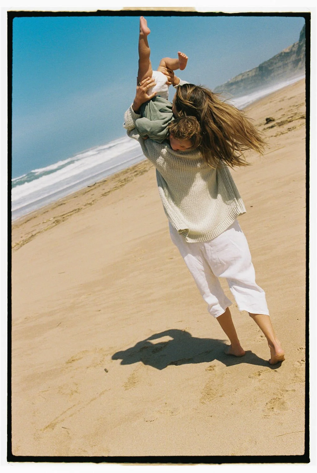 Keiarna from Gentle Currents with long hair wearing a cream sweater and white pants is holding her young boy upside down on a sandy beach with cliffs and the ocean in the background.