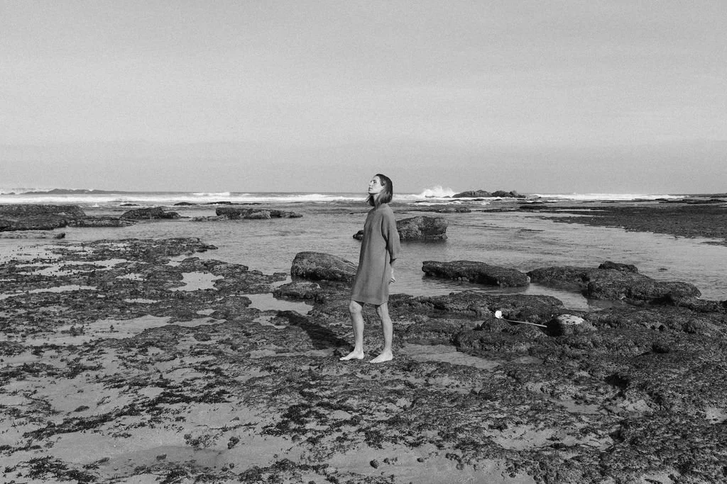 Keiarna from Gentle Currents standing on a rocky beach near the ocean, looking up with her eyes closed, wearing a loose dress.