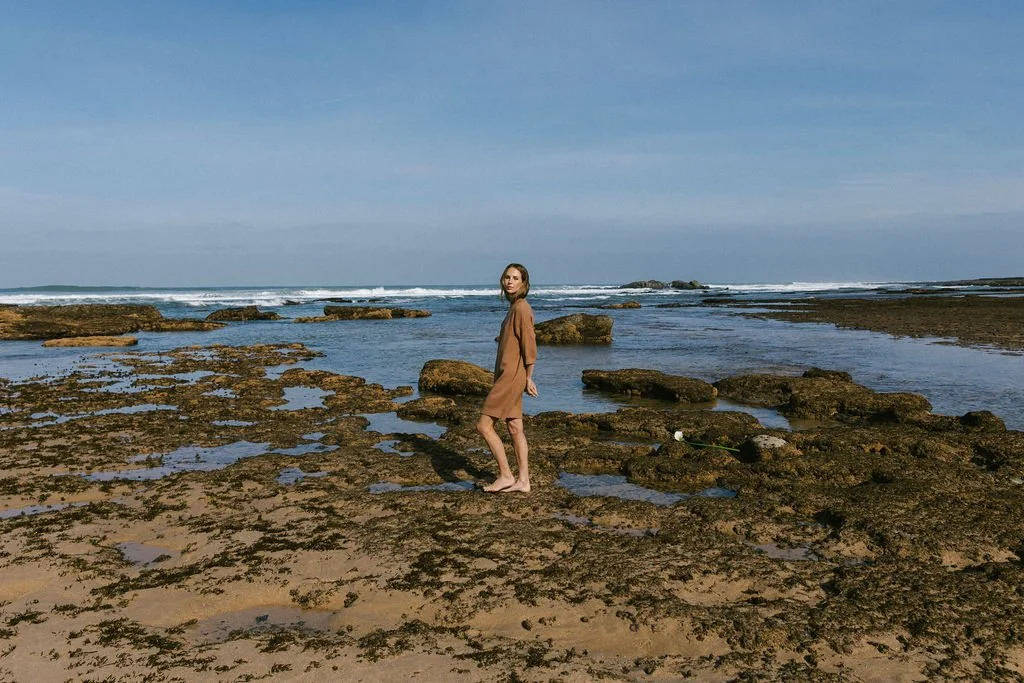 Keiarna from Gentle Currents in a tan dress walking barefoot on a rocky beach with tide pools, ocean waves, and a clear blue sky in the background.