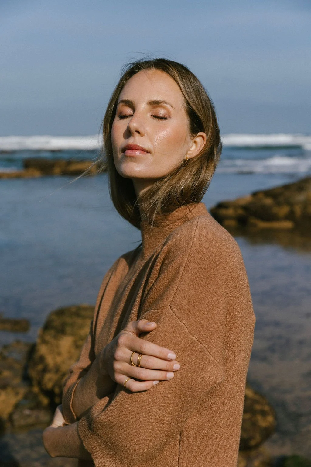 Keiarna from Gentle Currents with shoulder-length brown hair standing outdoors near the ocean, with rocks in the background. She has her eyes closed and is wearing a brown sweater, with her arms crossed, and appears to be enjoying the moment.