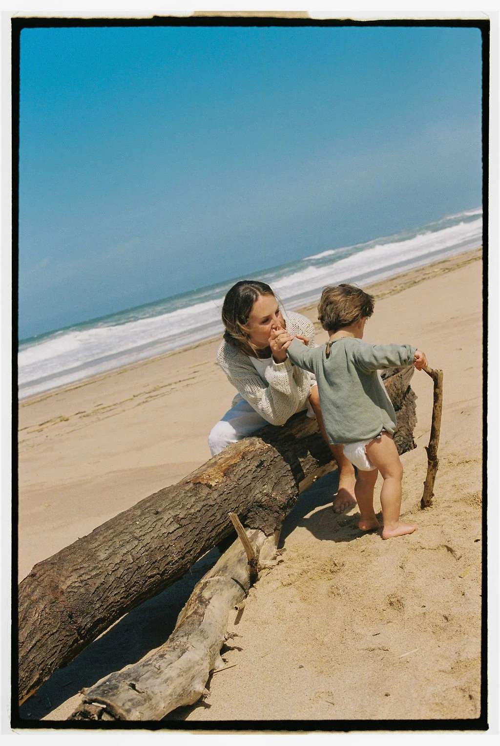 Keiarna from Gentle Currents and her young child sit on a large piece of driftwood on a sandy beach, with ocean waves and a clear blue sky in the background. The woman is holding the child's hand and appears to be whispering or talking to the child.