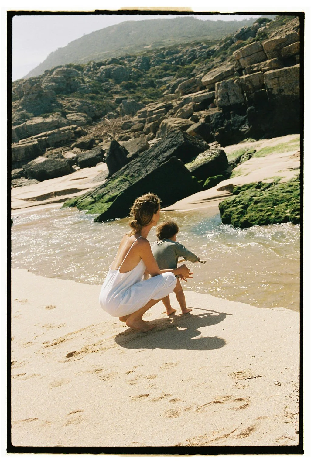 Keiarna from Gentle Currents and her child are at the beach, with the woman crouching and watching the child who is reaching into the water, surrounded by rocks and a hillside in the background.