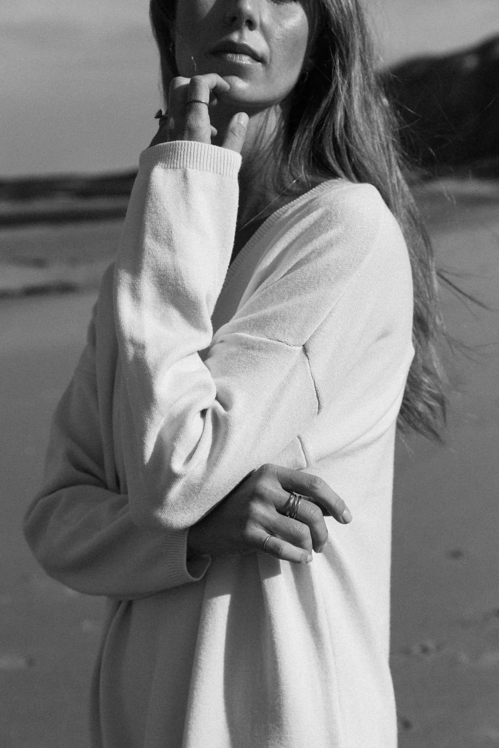 Black and white photo of Keiarna from Gentle Currents wearing a sweatshirt, standing outdoors with beach and ocean in the background, touching her face and chin with her hand.