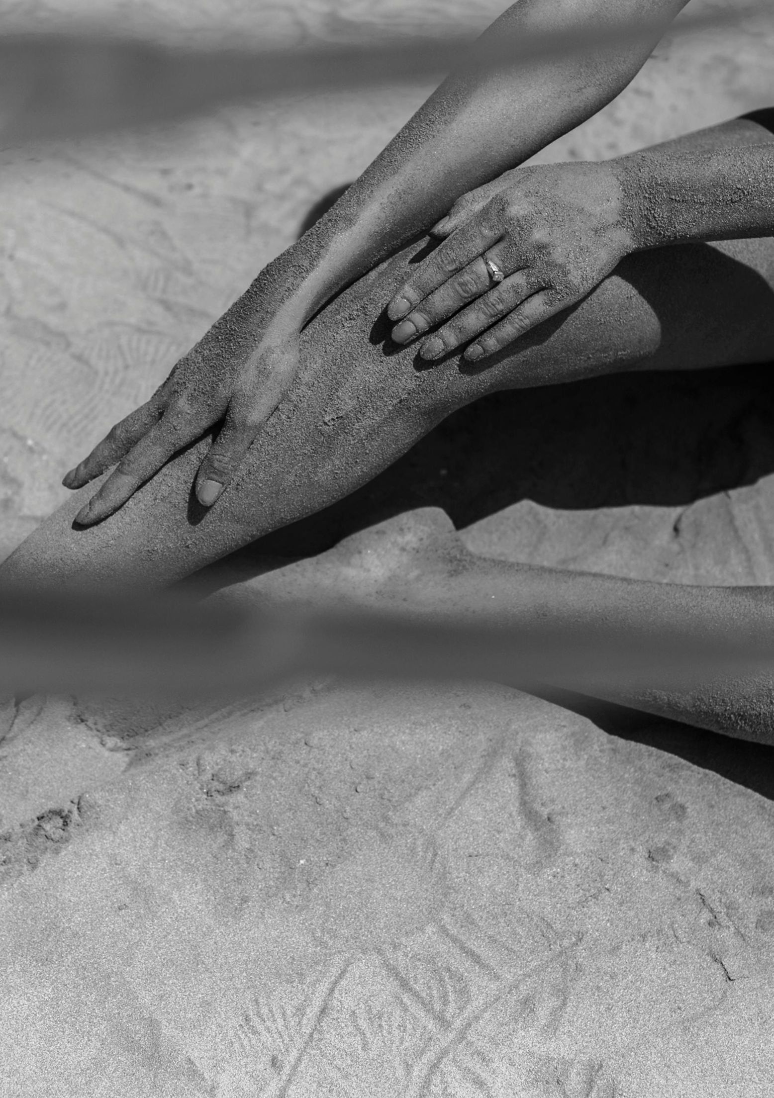 Black and white photo of a person's hand with a ring resting on their knee, on a sandy beach with footprints in the sand.