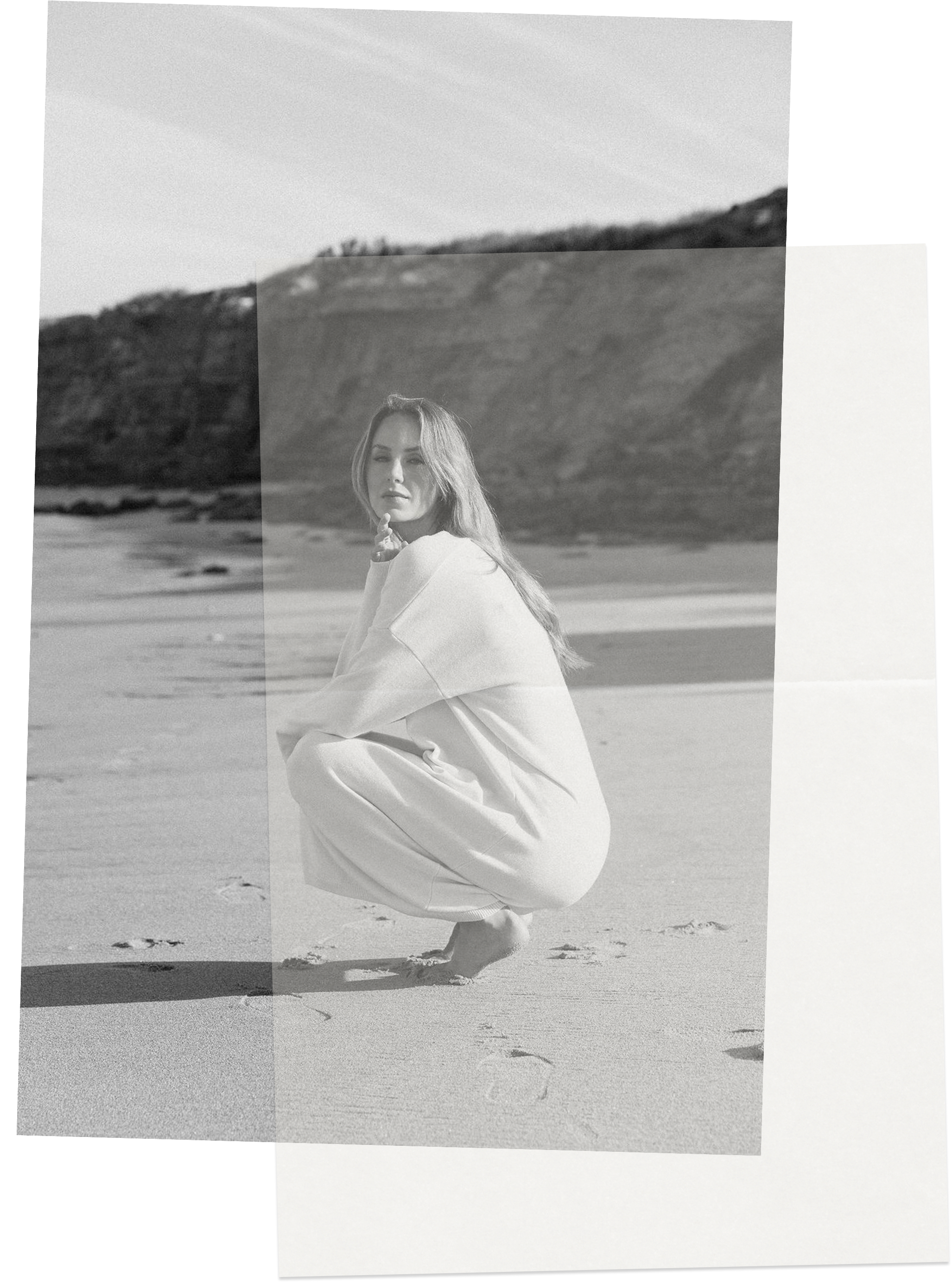 Black and white photo of Keiarna from Gentle Currents in white clothing crouching on a beach with the ocean and a hill in the background.