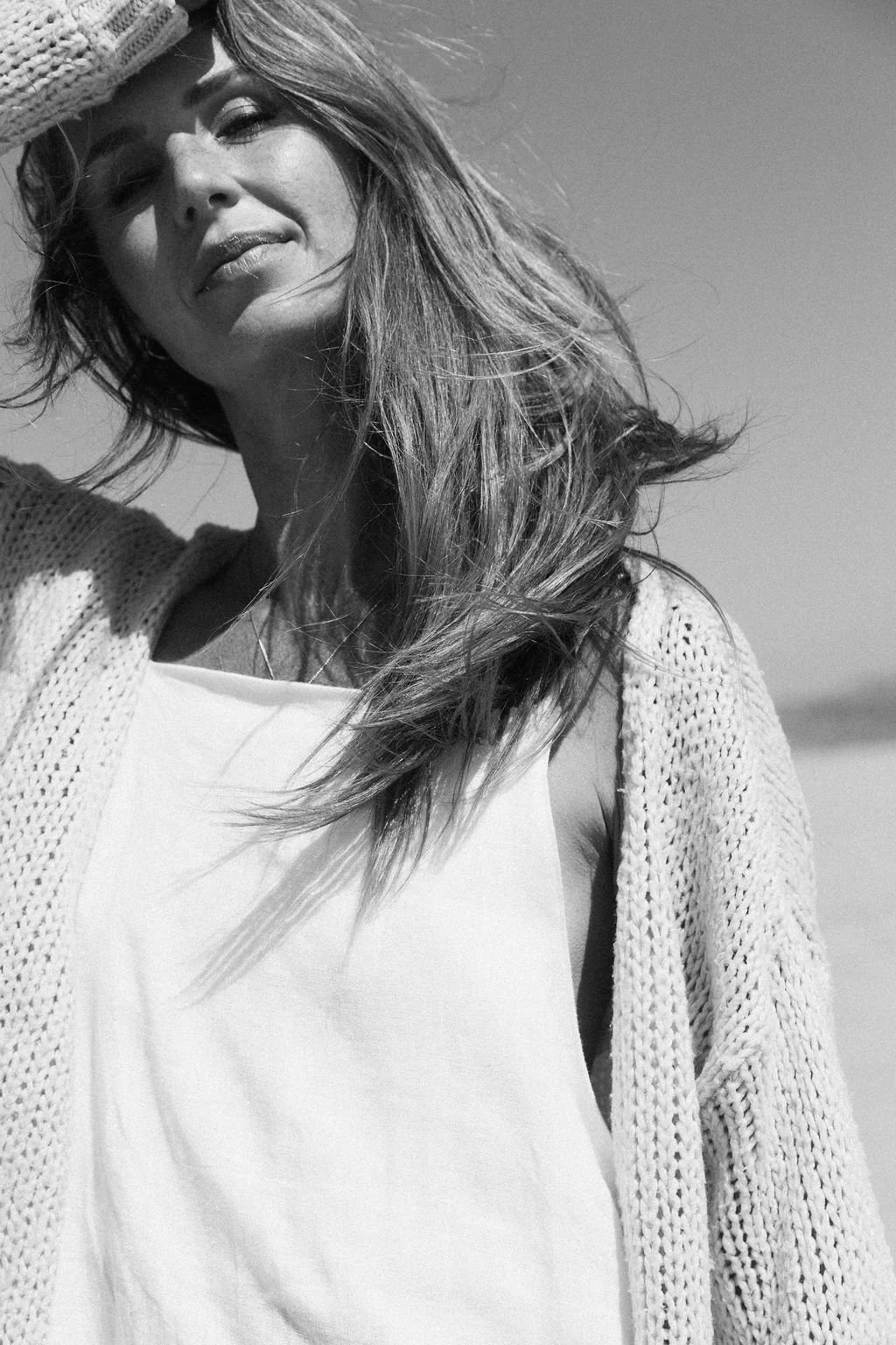 Black and white photo of a Keiarna from Gentle Currents with long hair, smiling slightly, shielding her eyes from the sun with her hand, wearing a knitted cardigan over a plain top, outdoors.