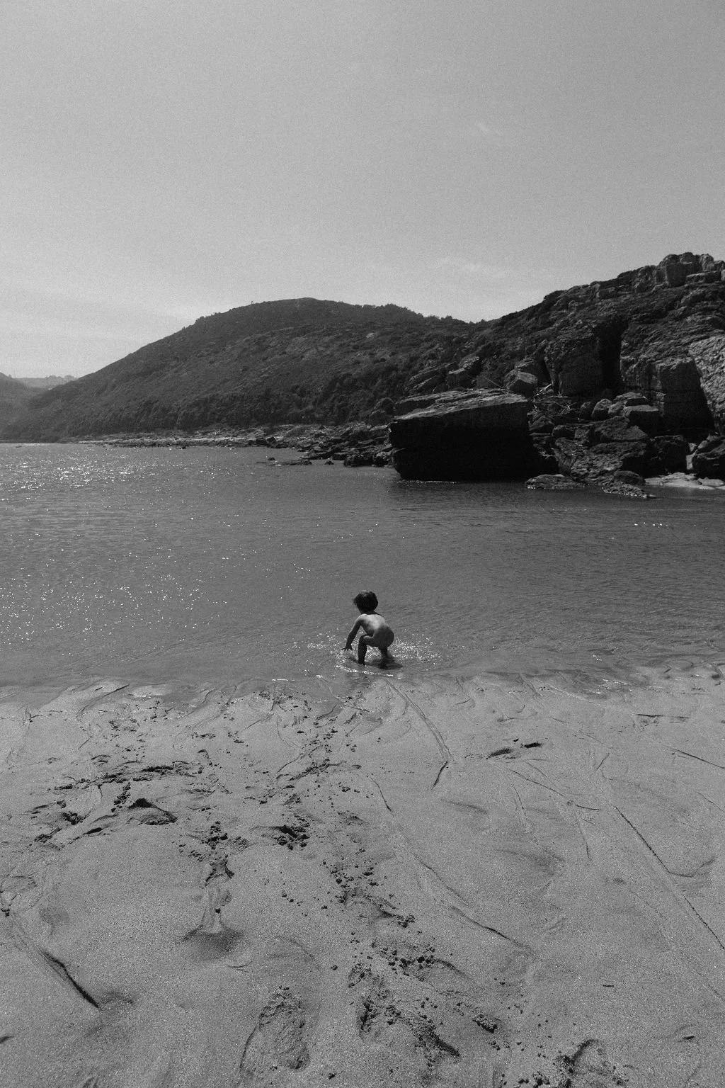 A young child playing in shallow water at a beach, with rocky cliffs and hills in the background, black and white photograph.