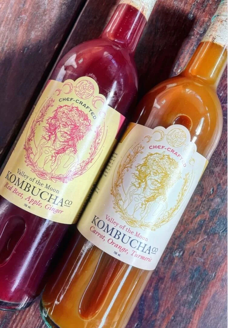 Two bottles of kombucha on a wooden surface, one with a red liquid and one with an orange liquid. Their labels feature a lion illustration and the text "Valley of the Moon Kombucha Co."