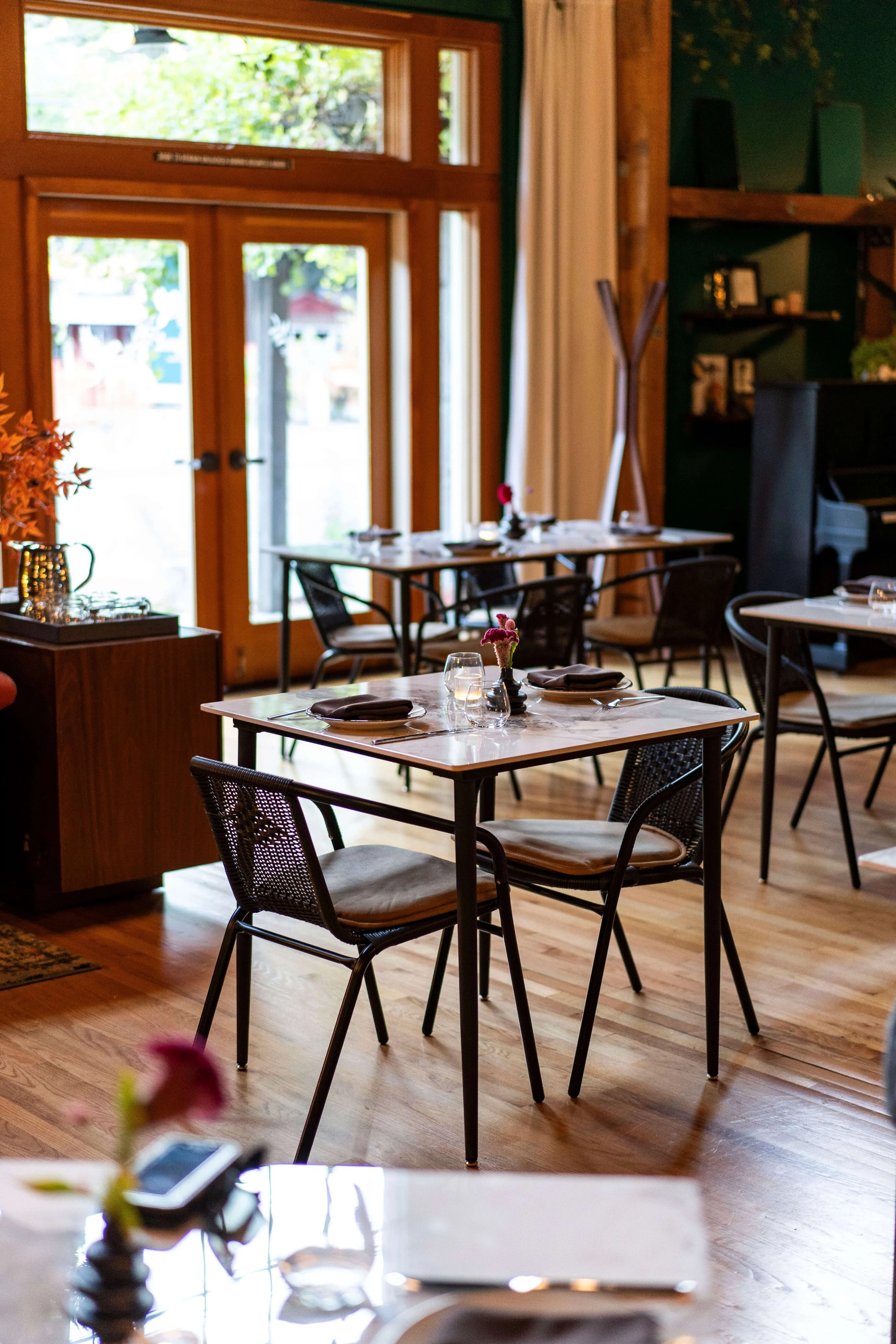 Interior of a cozy restaurant with wooden floors, large windows, and simple, modern tables set with napkins, glasses, and small flower centerpieces.