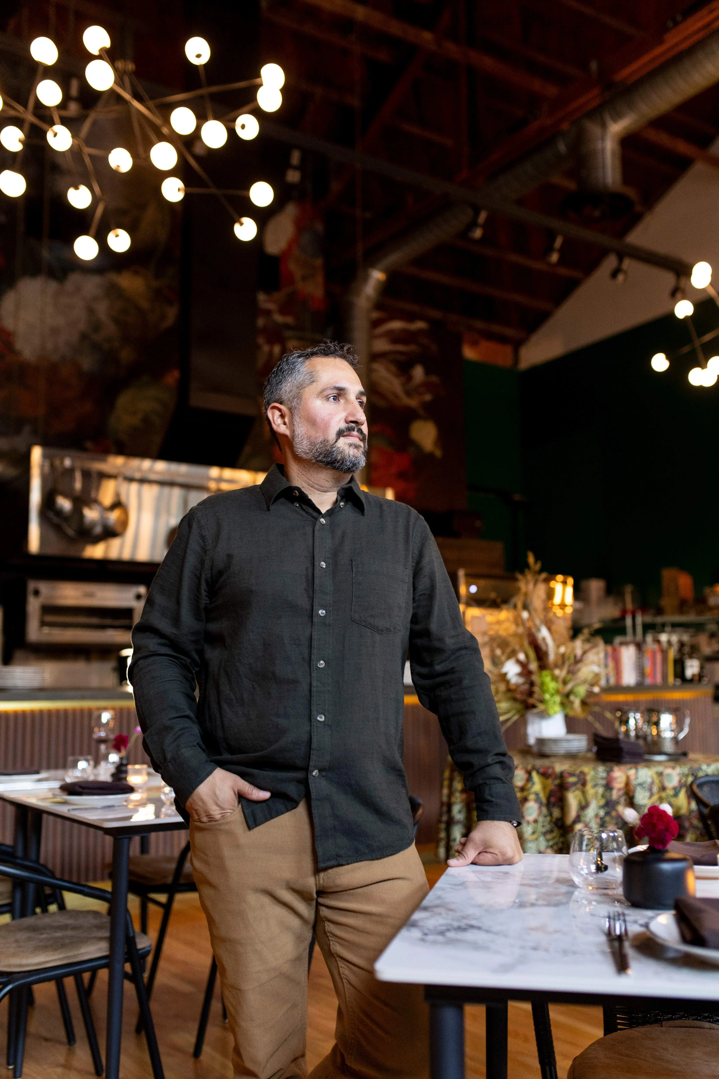 A man with short gray hair and beard, wearing a black button-up shirt and khaki pants, stands at a table in a restaurant with a modern industrial design, decorated with flowers and table settings. The background features warm lighting, artwork, and kitchen equipment.