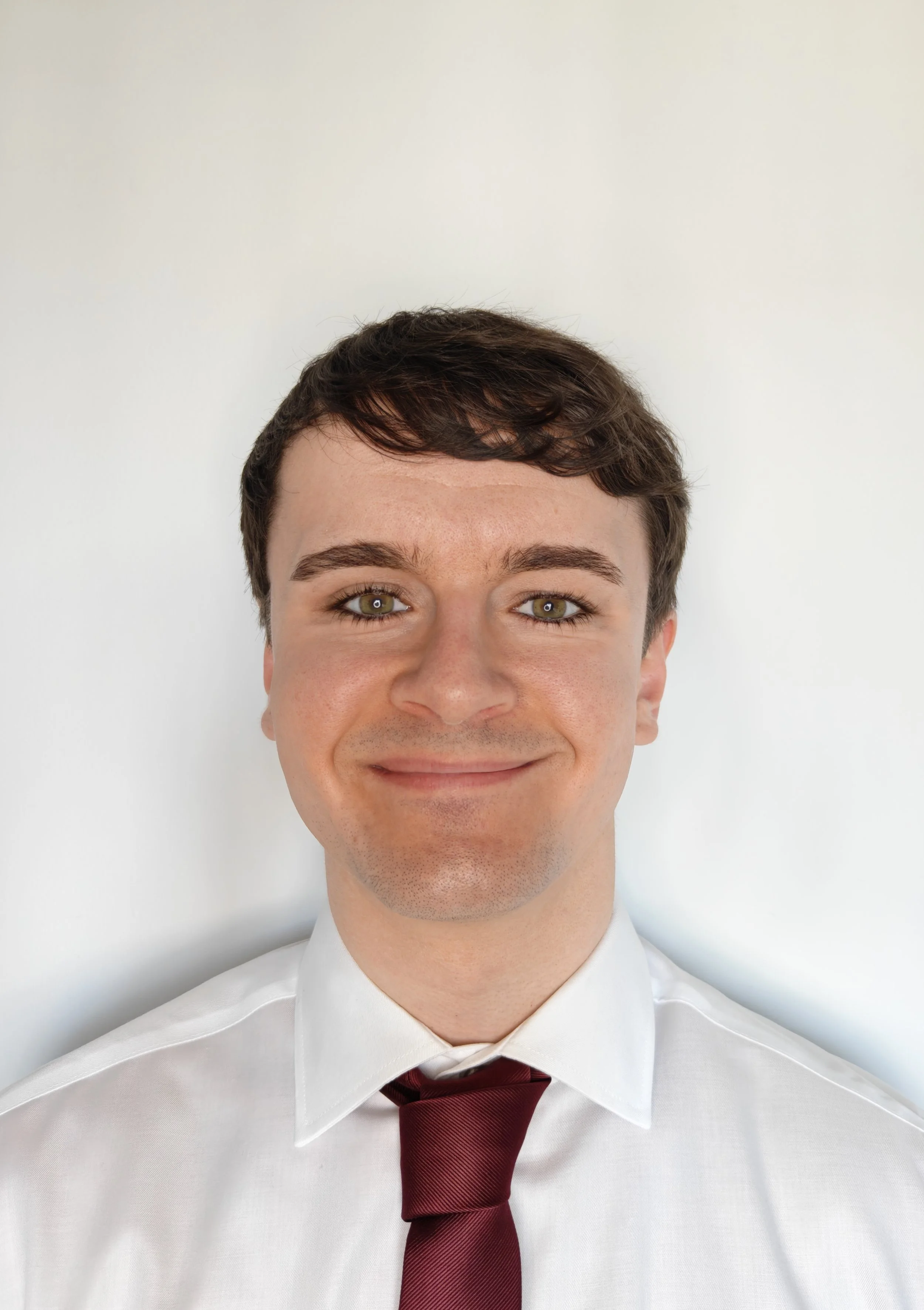 Retrato de un hombre joven con cabello castaño oscuro, ojos verdes, sonriendo, vestido con camisa blanca y corbata burdeos, fondo blanco.