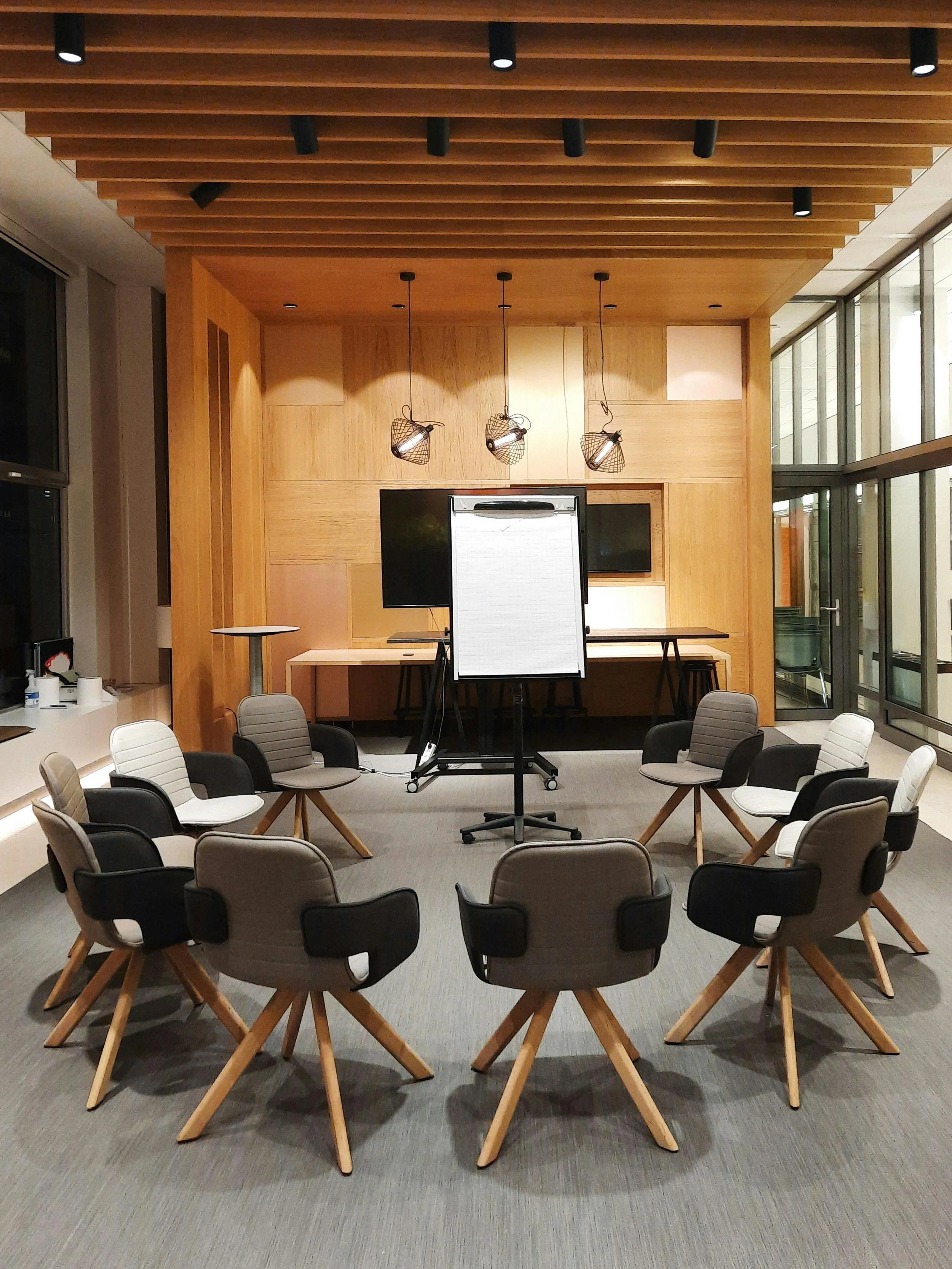 Sala de reuniones con sillas negras y blancas alrededor de una mesa de madera. En el fondo, una pared de madera con iluminación, y en el centro, un pizarrón en blanco en un soporte móvil. Techos de madera y ventanas grandes con vista exterior.