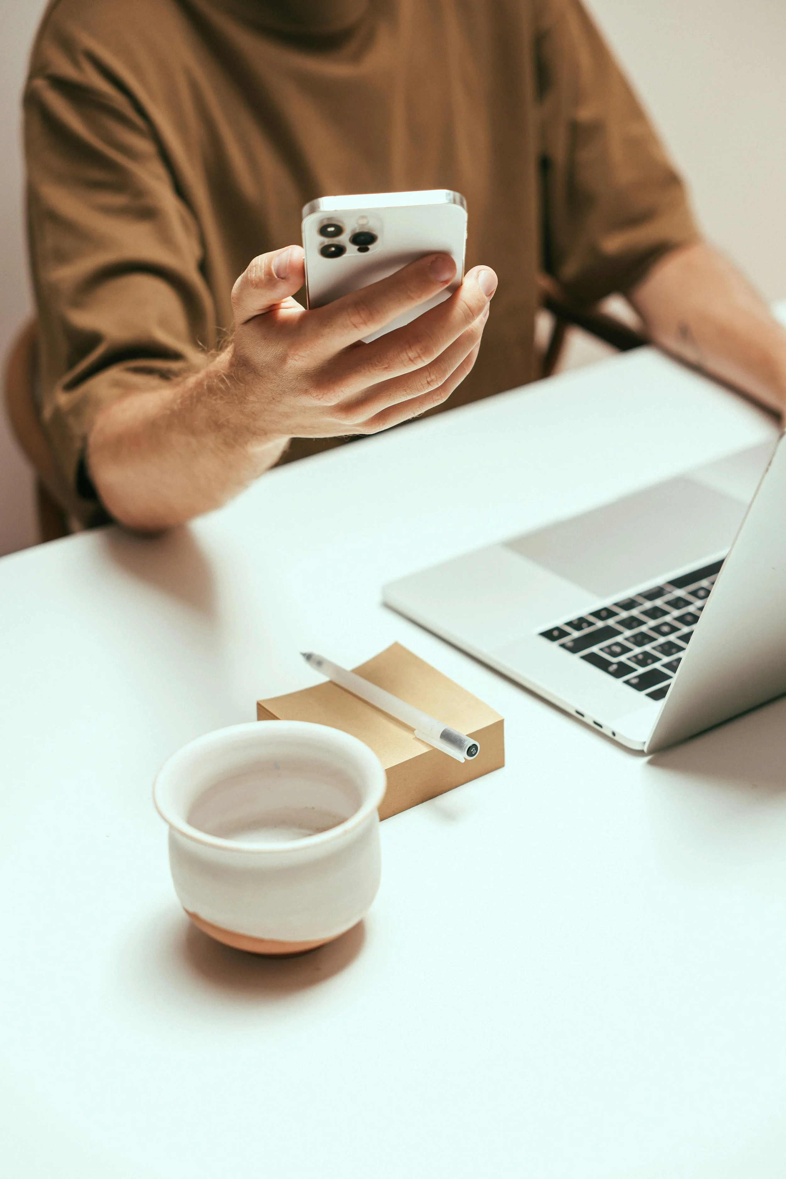 Persona usando un teléfono inteligente en una mesa blanca con una taza, un cuaderno, un bolígrafo y una computadora portátil.