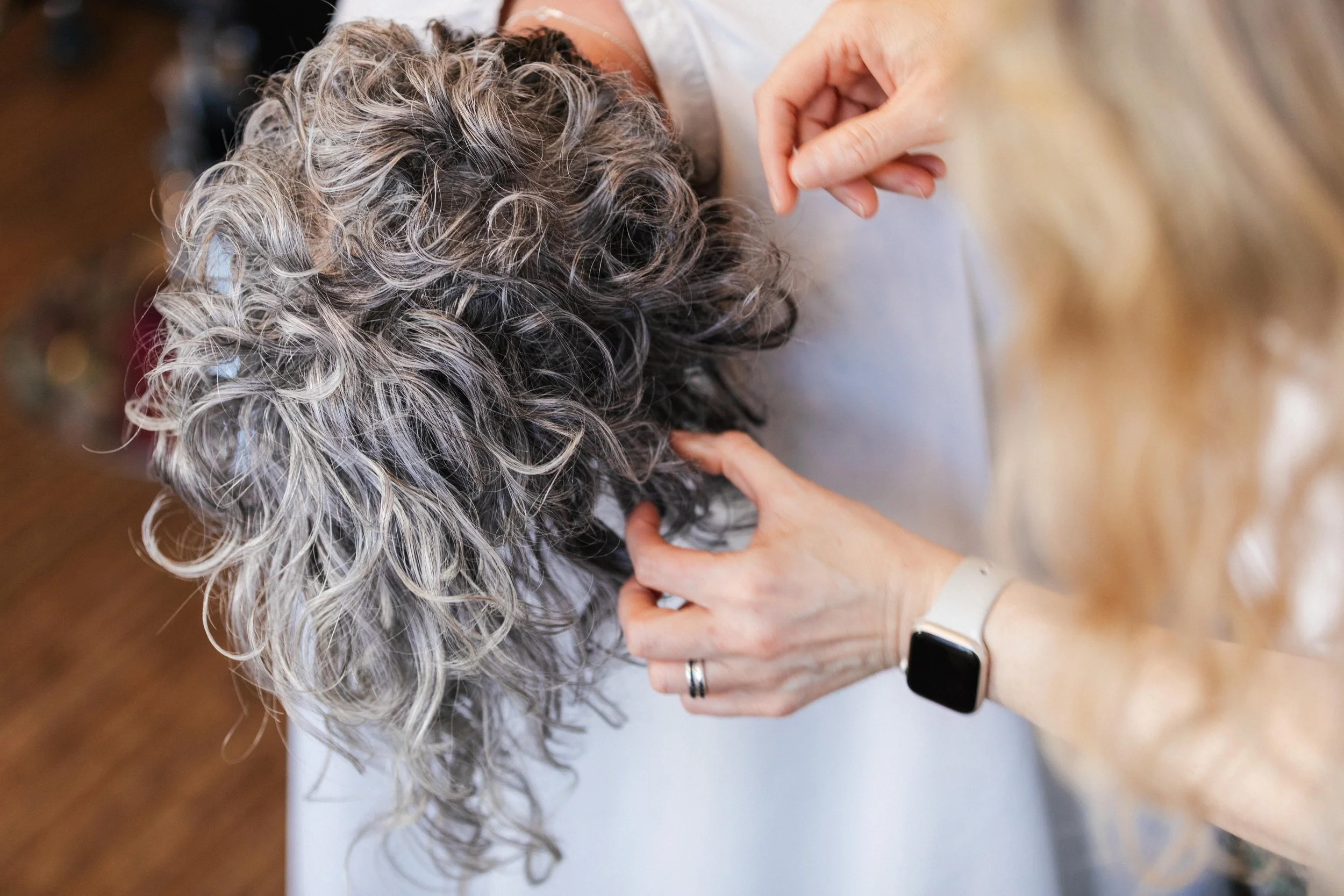 A person with curly gray hair is getting a haircut or styling at a salon. The stylist's hands are visible, working on the hair. The stylist is wearing a smartwatch and a ring.