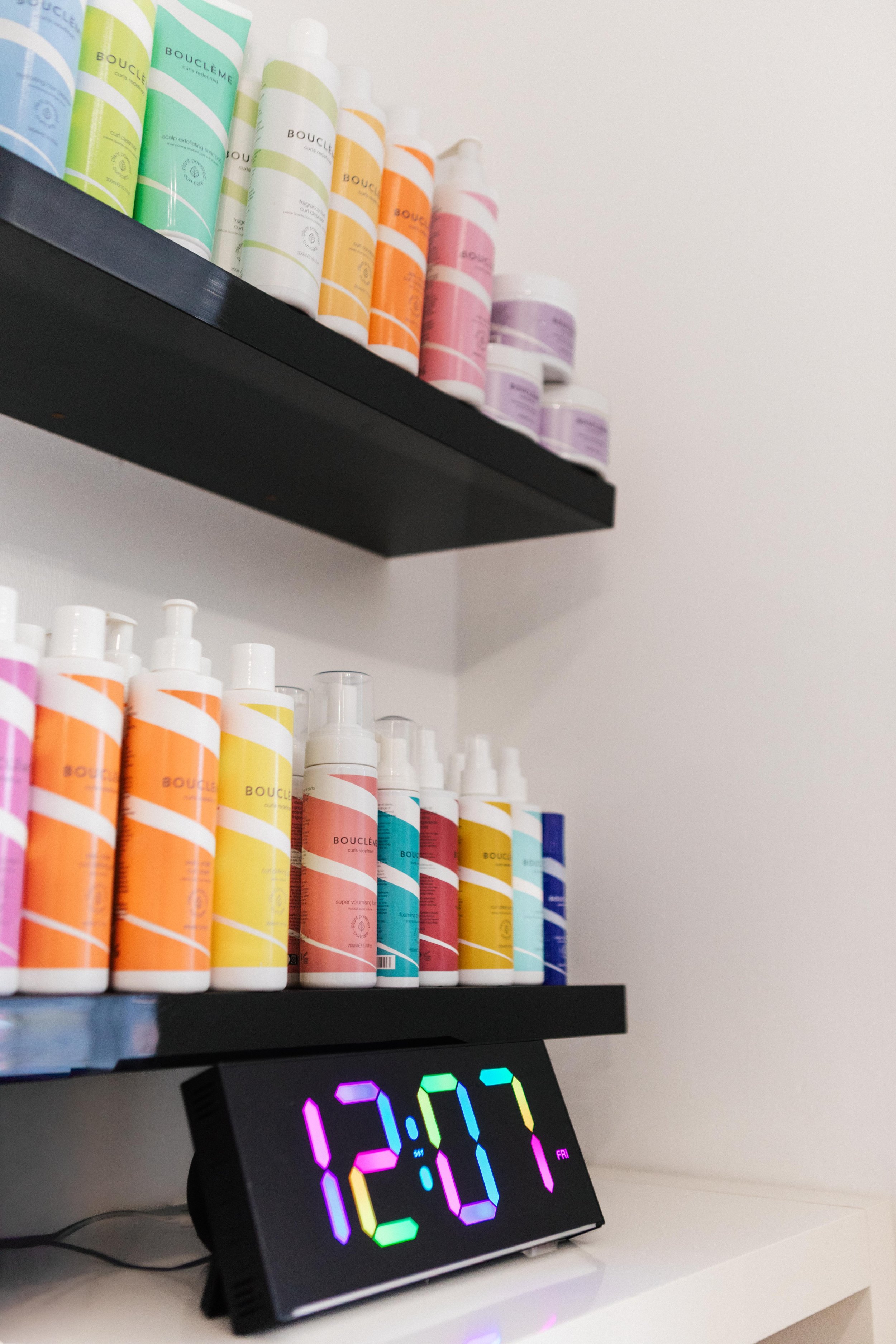 Colorful bottles of hair or skincare products on black shelves above a digital clock displaying 12:07.