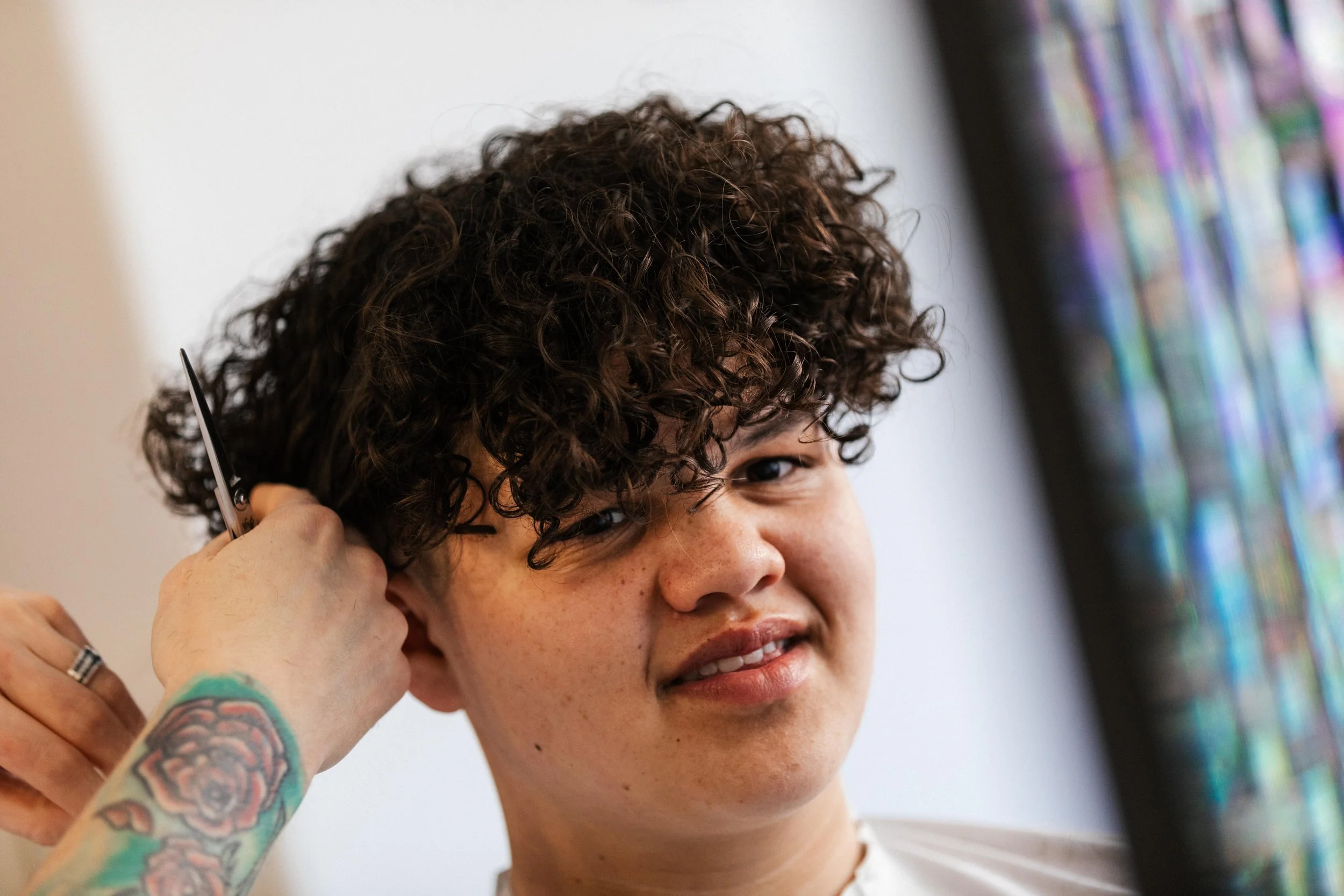 A person with curly dark hair getting a haircut, holding scissors near their ear, with tattoos on their arm, and a colorful display on a screen to the side.