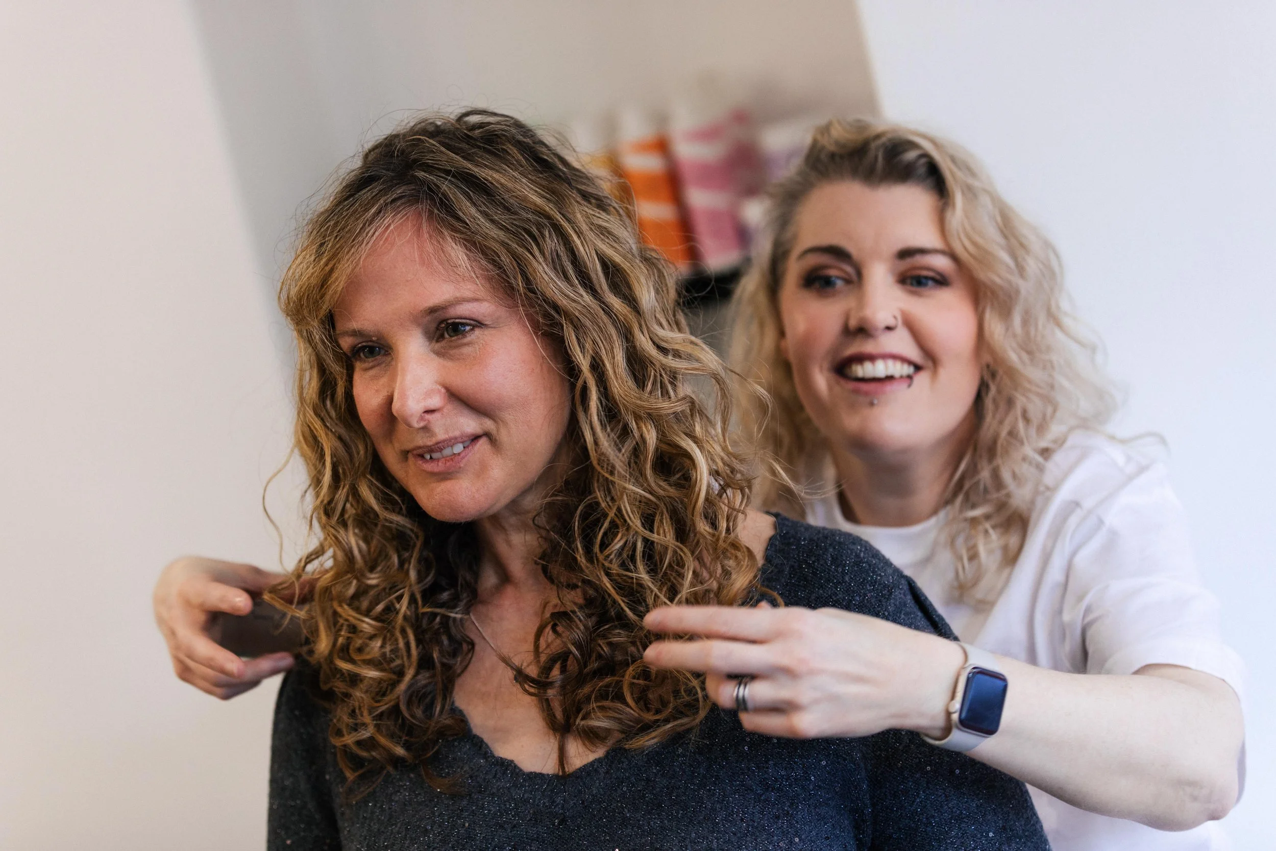 Two women, one with curly brown hair and the other with wavy blonde hair, are smiling as they style the brown-haired woman’s hair.