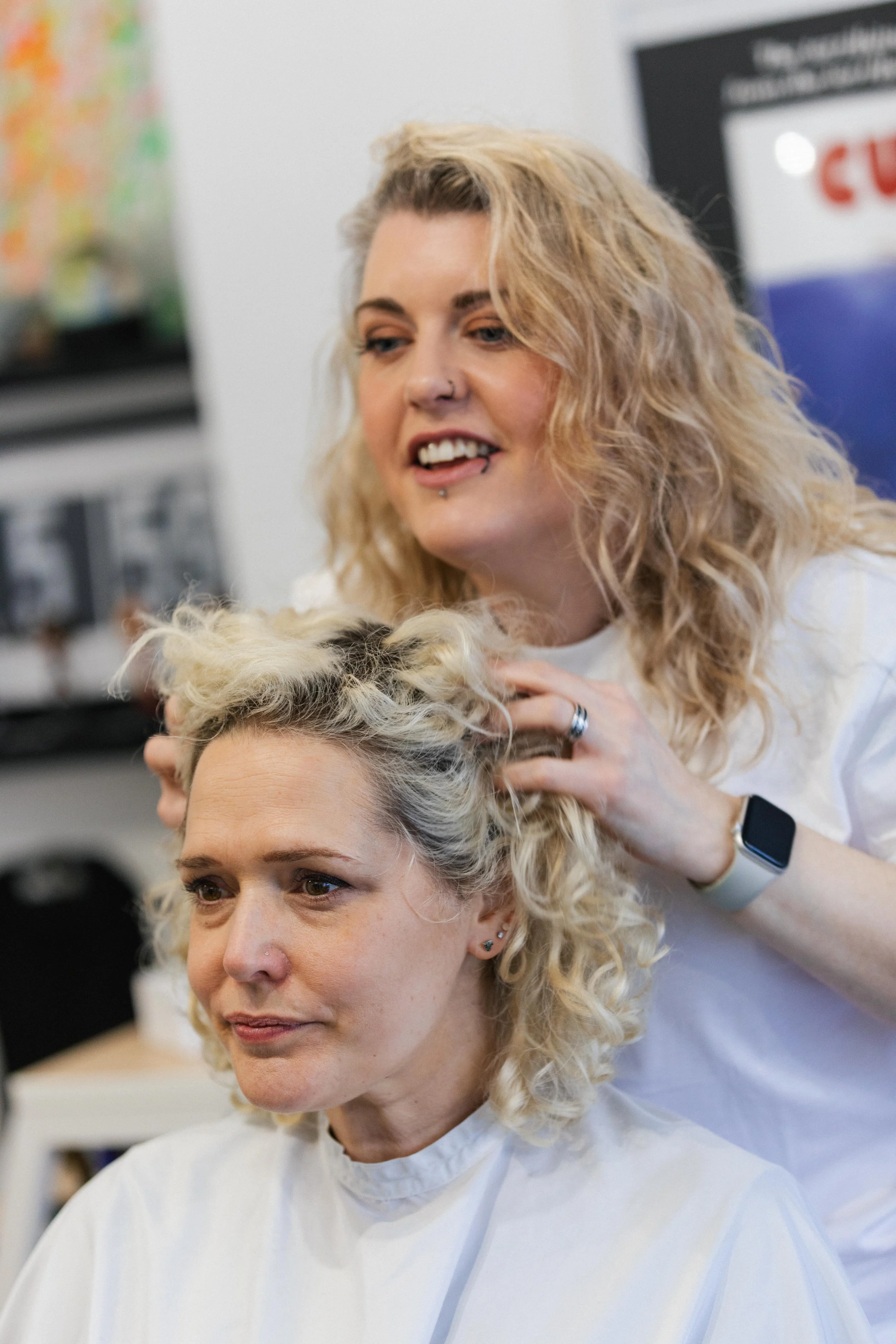 A hairstylist with curly blonde hair working on a woman's curly blonde hair in a salon.