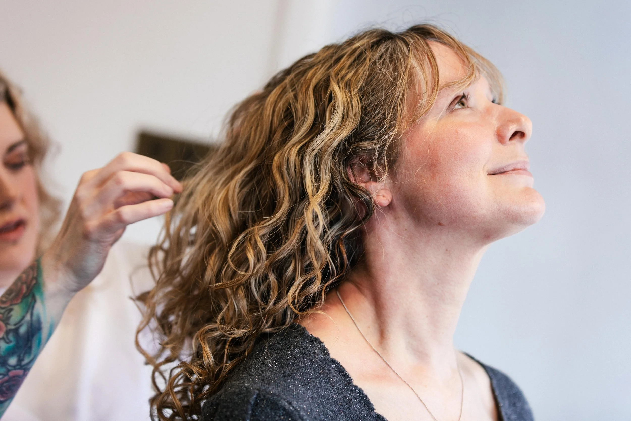 A woman with curly blonde hair getting her hair styled by a stylist with a colorful tattoo on her arm in a well-lit room.