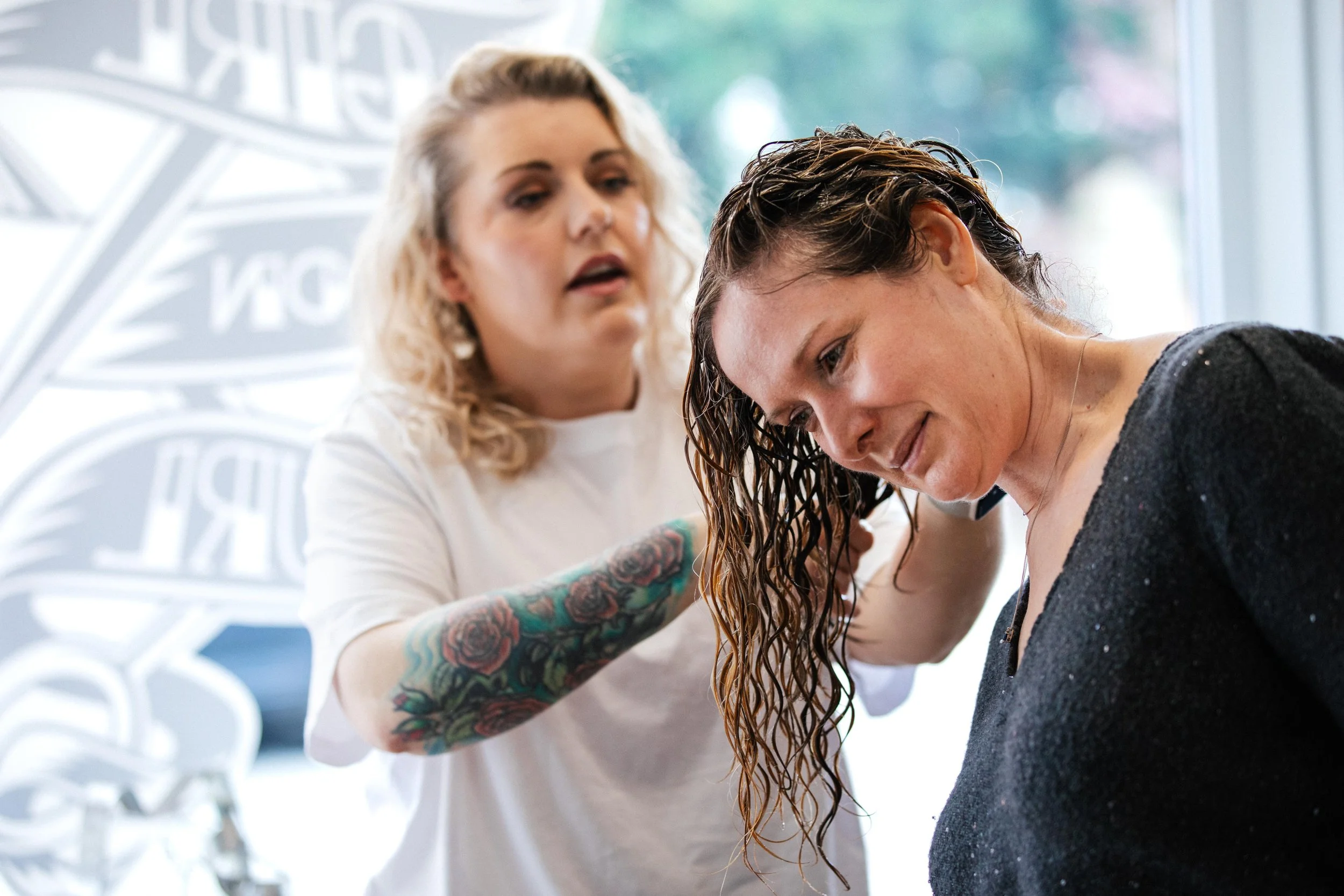 A hairstylist with a tattoo on her arm styling a woman's wet, curly hair in a salon.