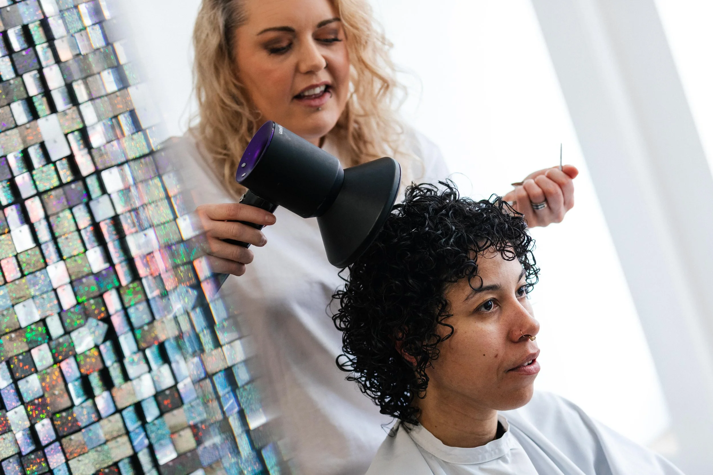 A woman getting her hair dried with a hairdryer in a beauty salon, with a stylist standing behind her holding a tool, and a holographic mosaic on the left side of the image.