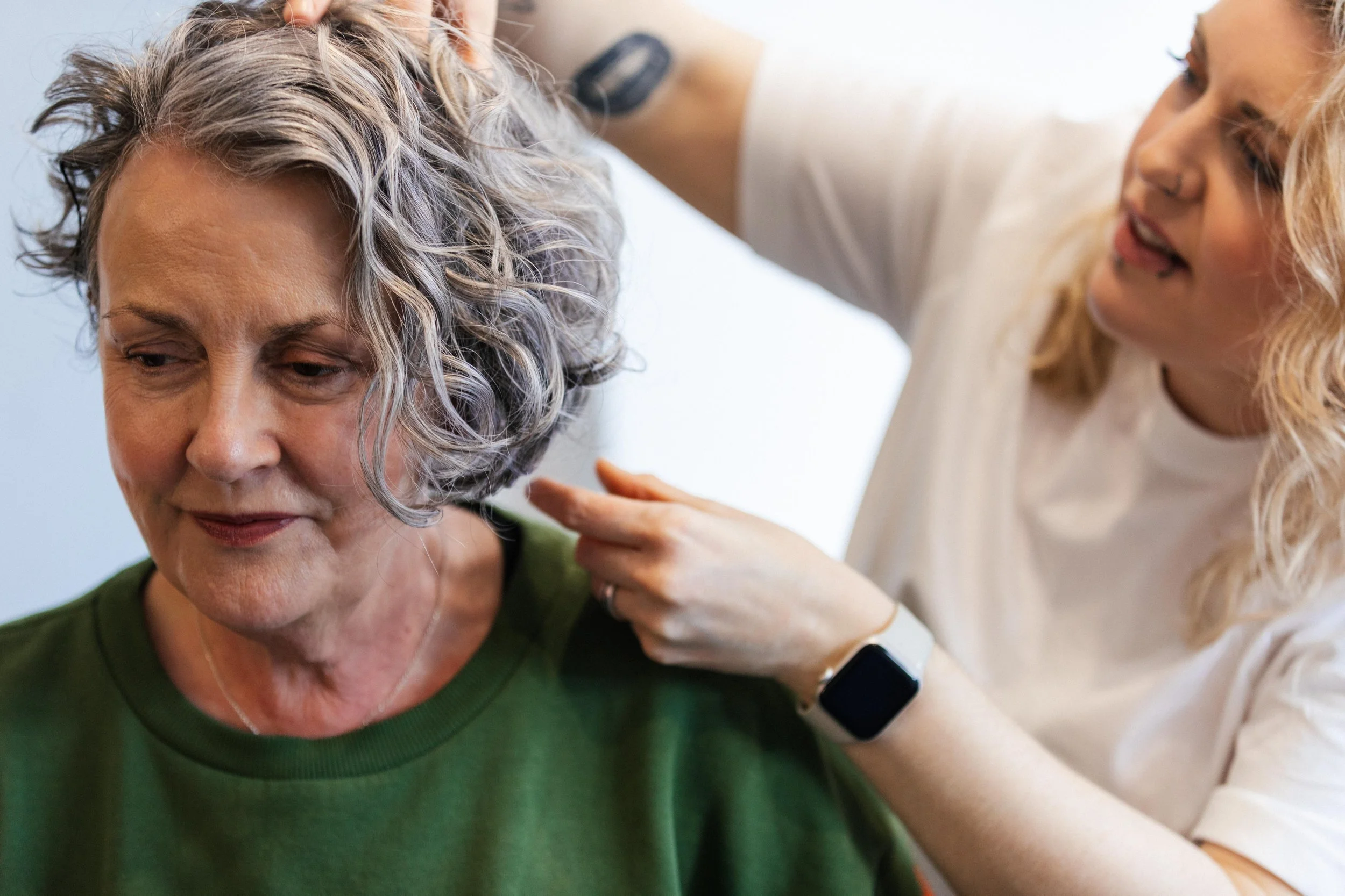 A woman with blonde curly hair is examining the scalp of an older woman with short gray curly hair. The older woman is wearing a green shirt and looking downward, while the younger woman, dressed in white, appears to be inspecting or providing some k