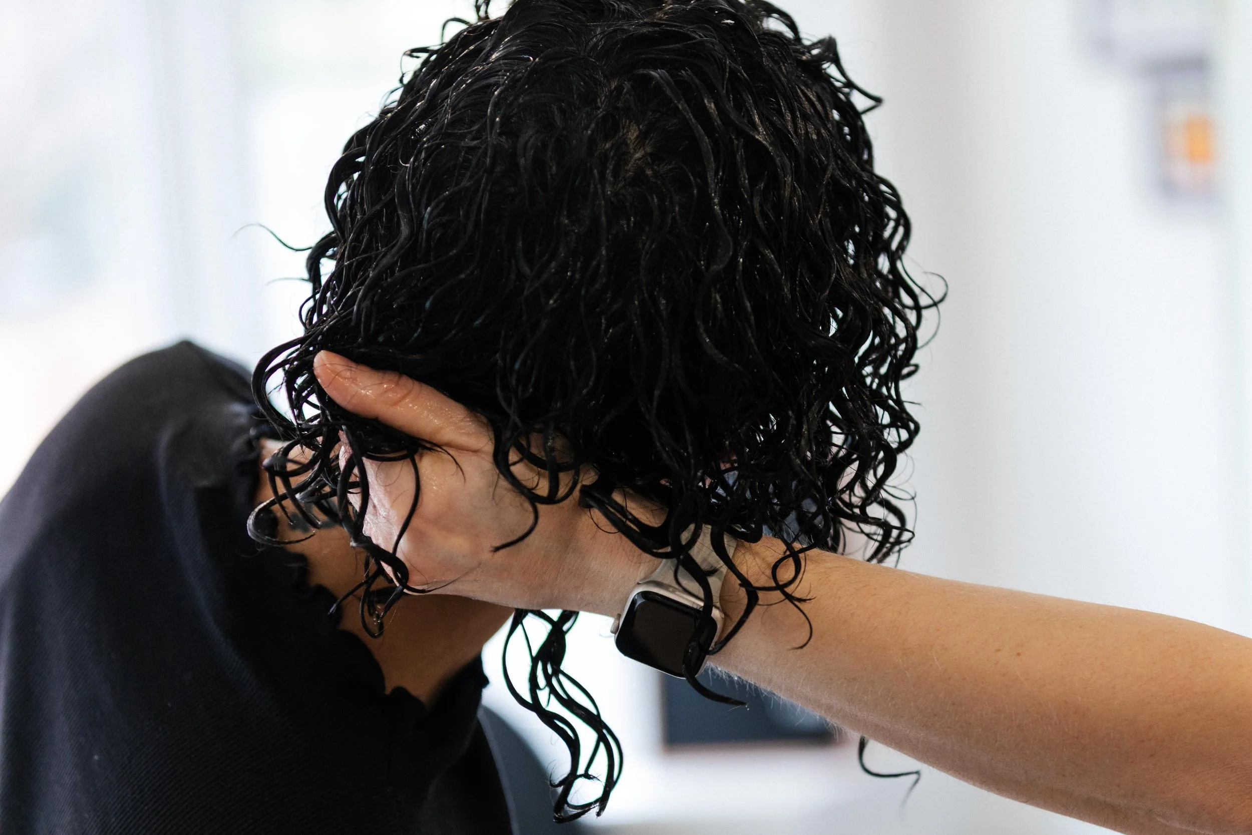 Person with wet, curly black hair holding their head with one hand, wearing a black shirt and a black smartwatch.