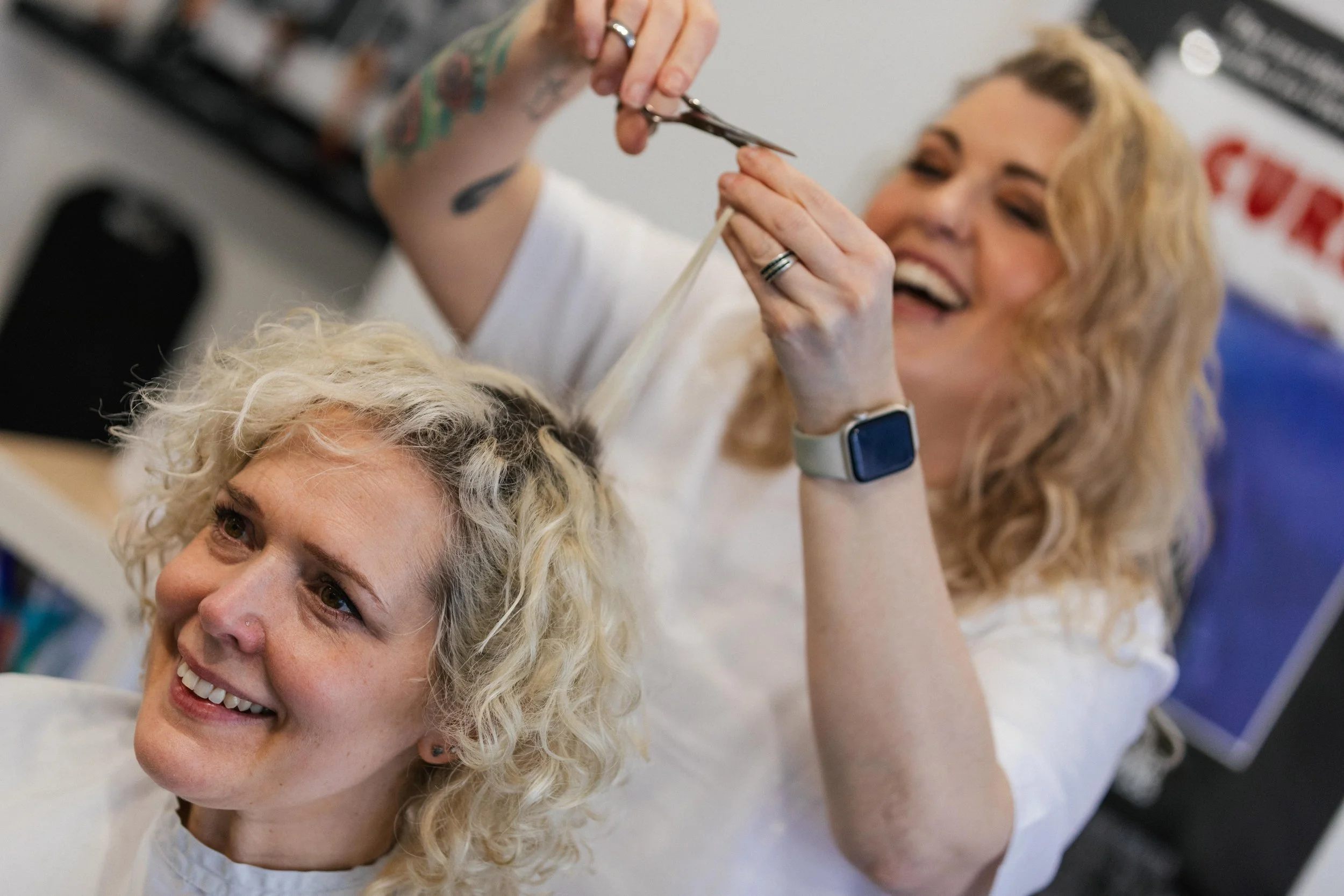 A hairstylist cutting a woman's curly blonde hair in a salon, both are smiling and happy.