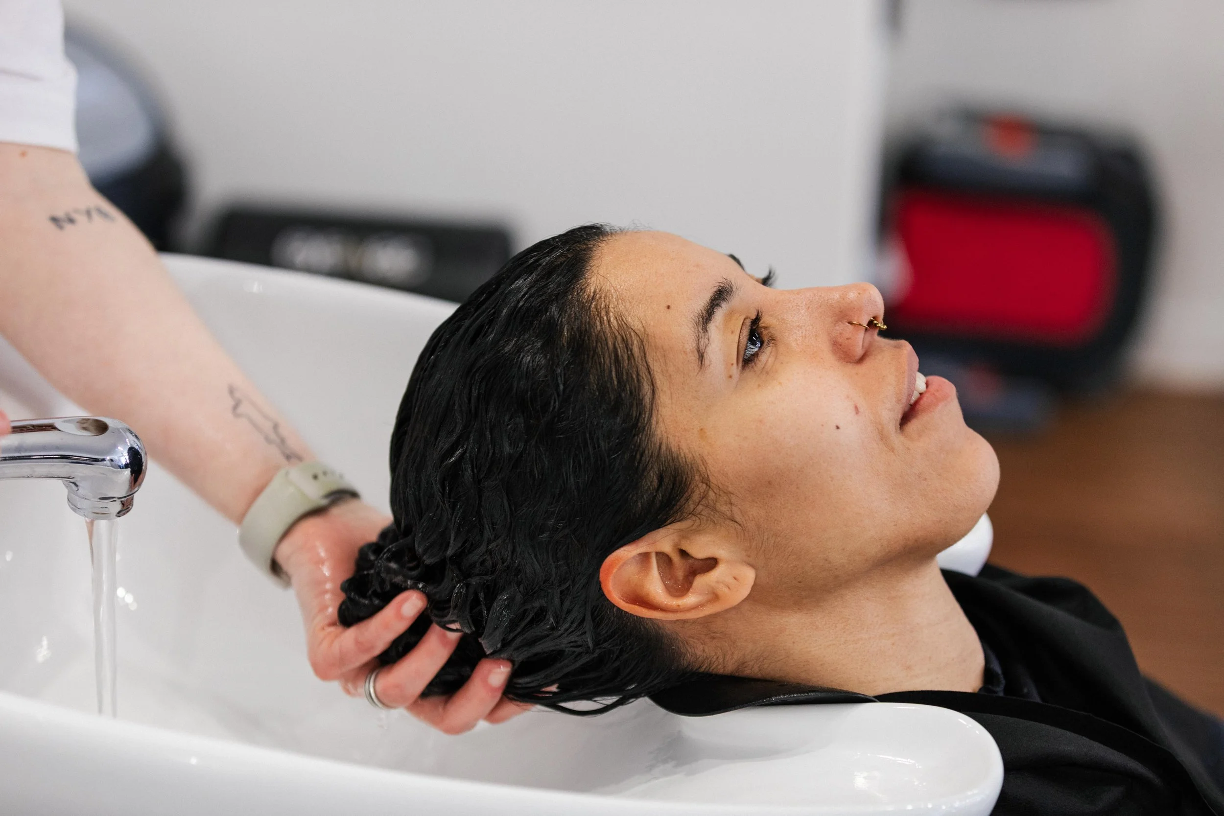 A woman with dark, wet hair receiving a head wash at a salon or barber shop, with her head leaning back in a sink as water is rinsed through her hair.